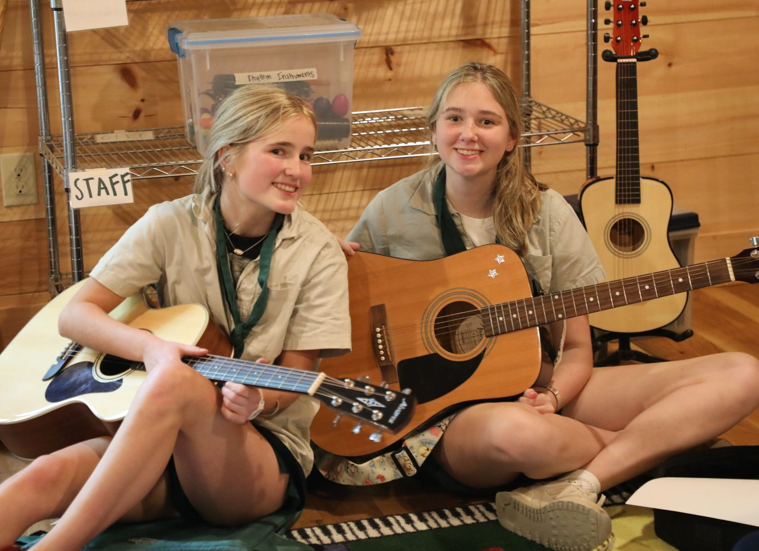 Two people smiling while sitting on the floor and playing acoustic guitars in a room with a staff sign and shelves.
