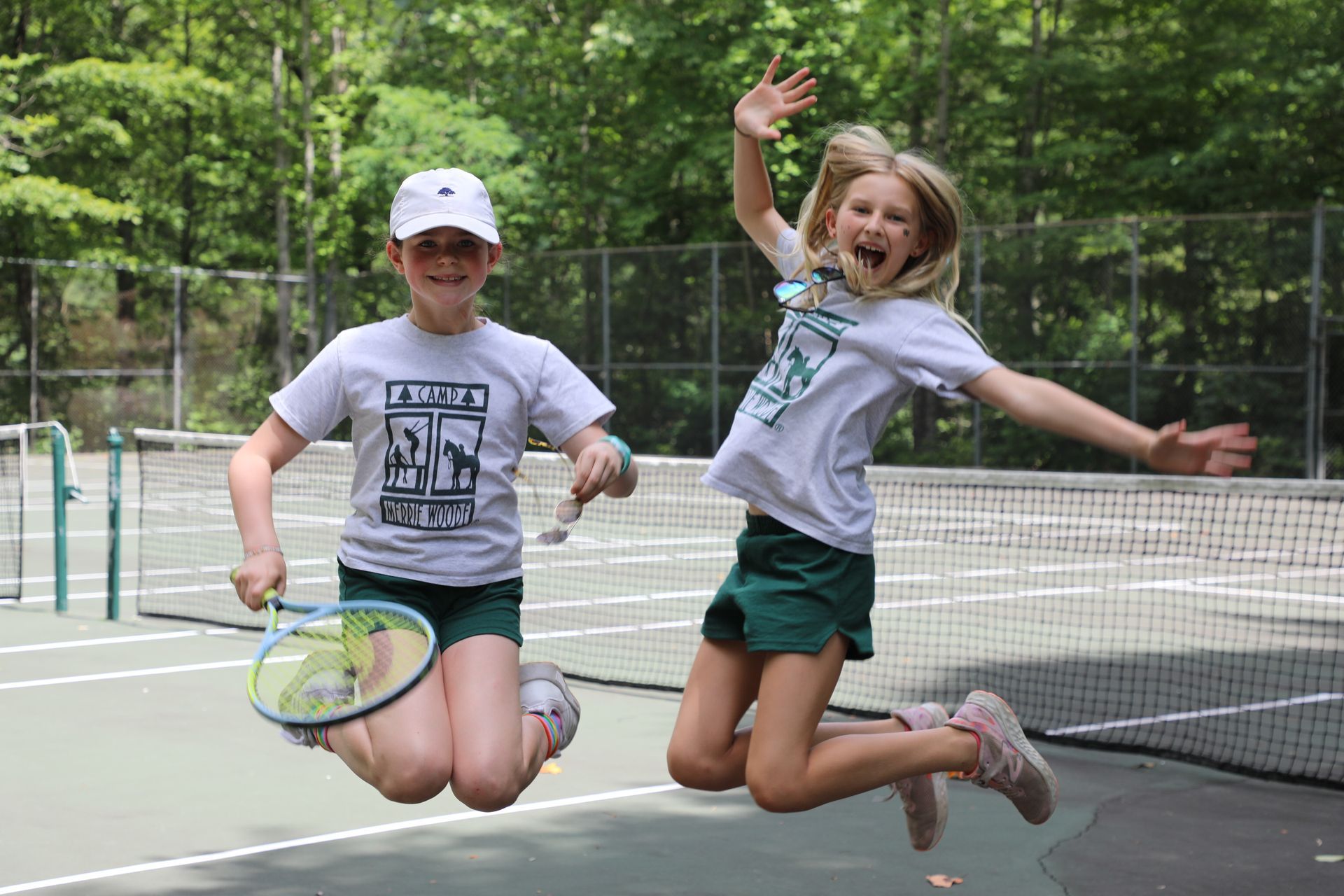 Two children in athletic clothes jump for joy on a tennis court, one holding a tennis racket.