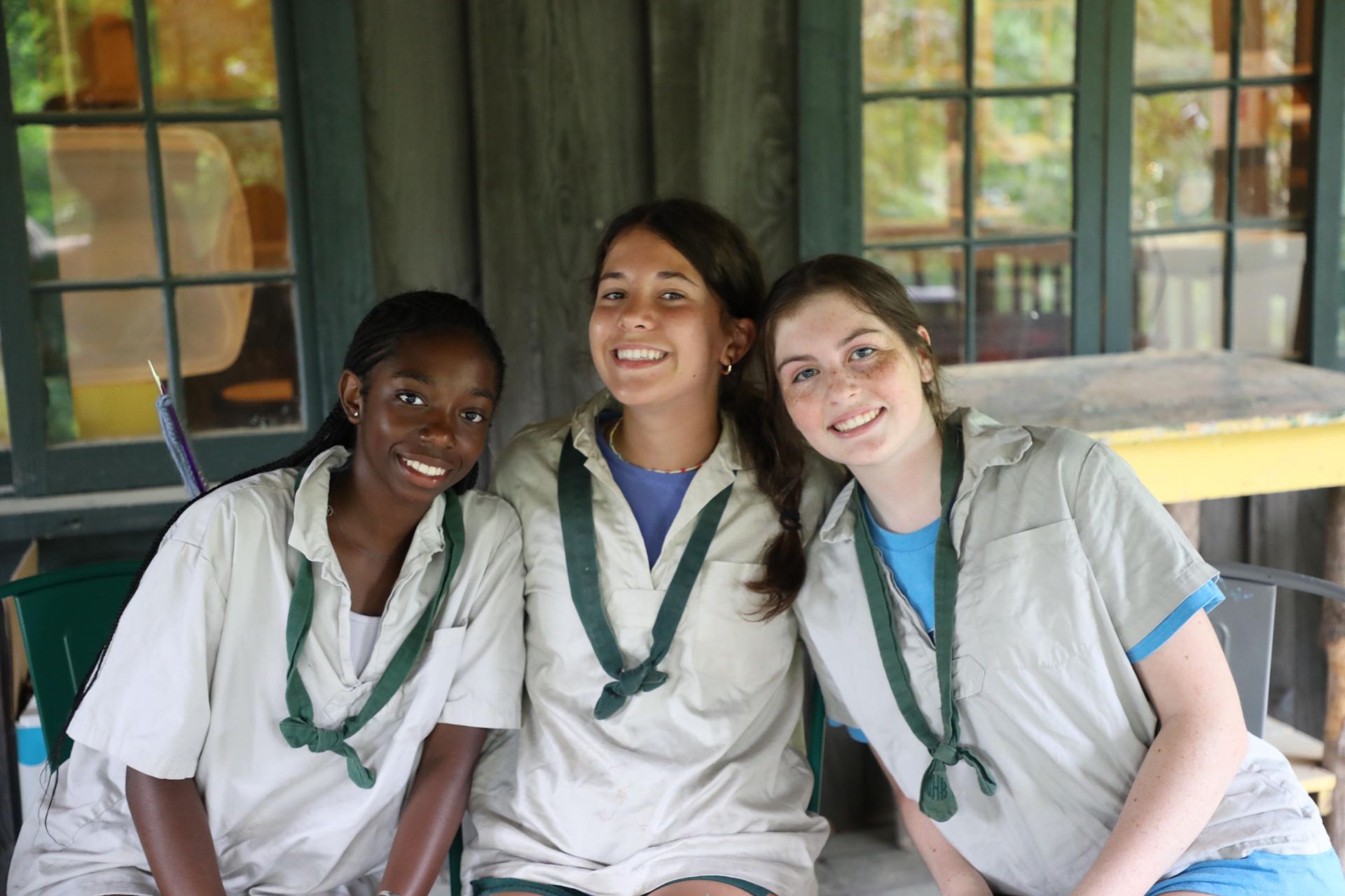 Three smiling people in beige shirts and green neckerchiefs pose together in front of a windowed building.