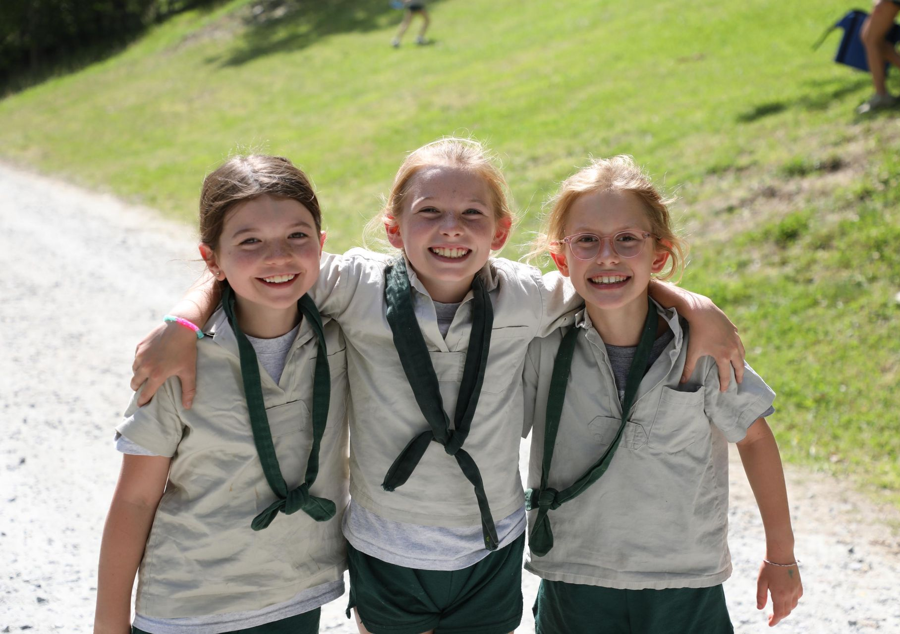 Three children in khaki scout uniforms with dark green scarves smile with arms around each other on a grassy hillside.