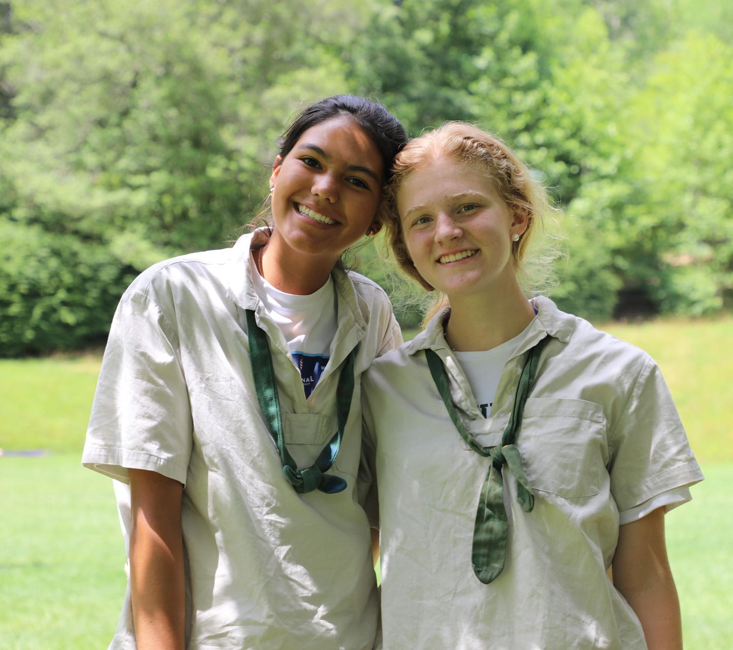 Two people in beige scout uniforms with green neckerchiefs smiling together in a grassy outdoor setting.