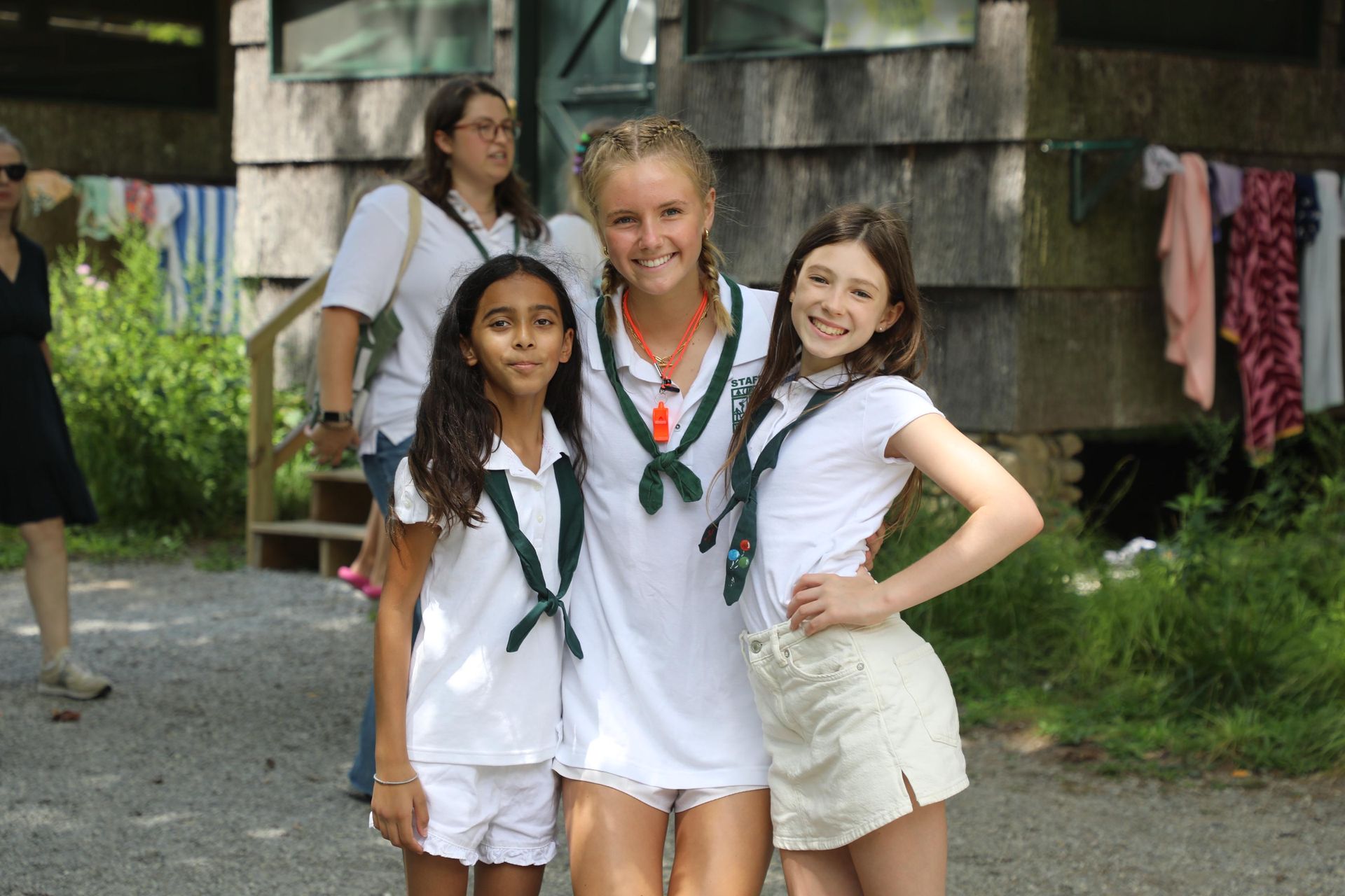 Three people in white shirts and dark scarves pose smiling in front of a rustic wooden building.
