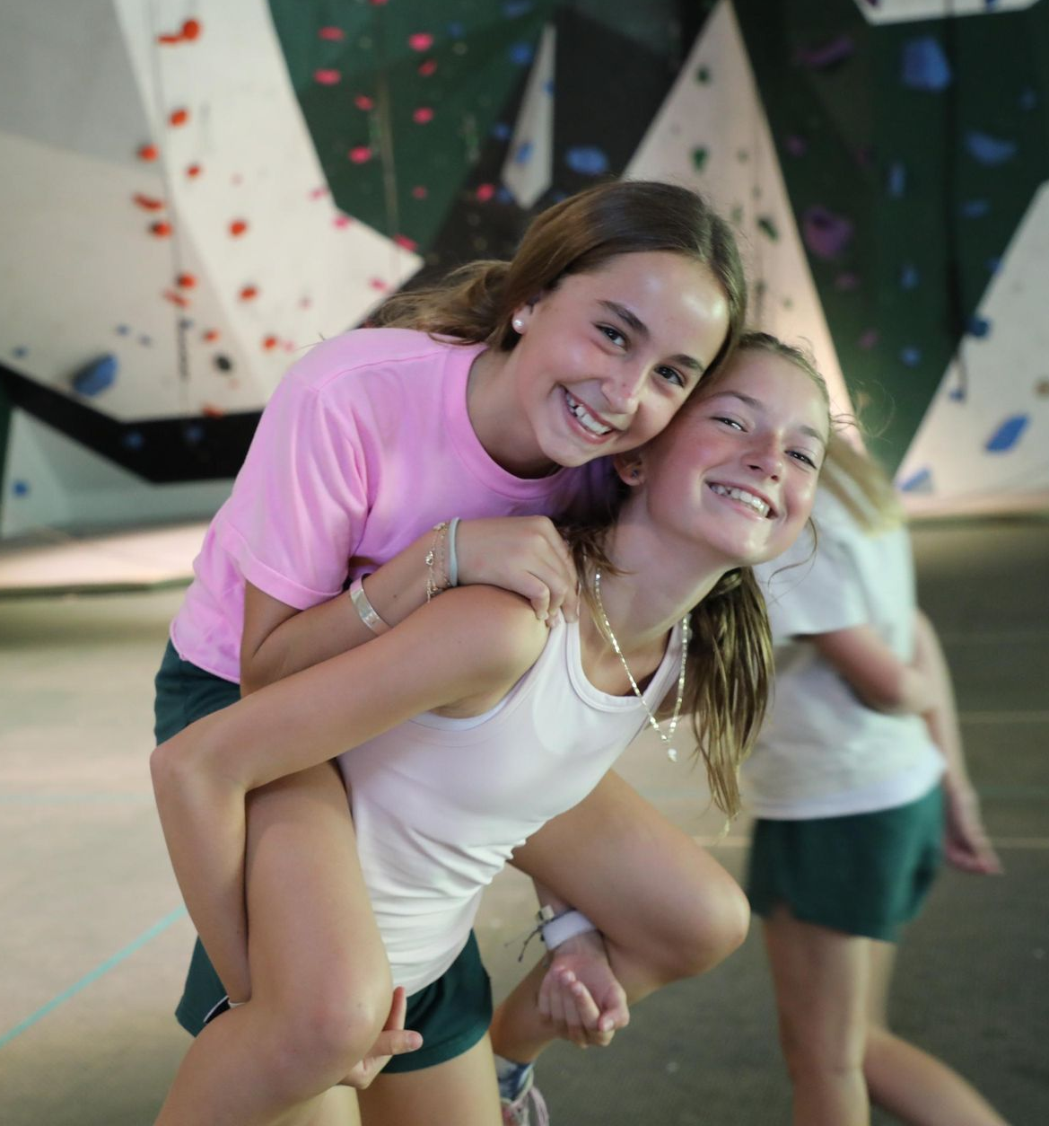 Two smiling people in a rock climbing gym, one giving the other a piggyback ride in front of climbing walls.