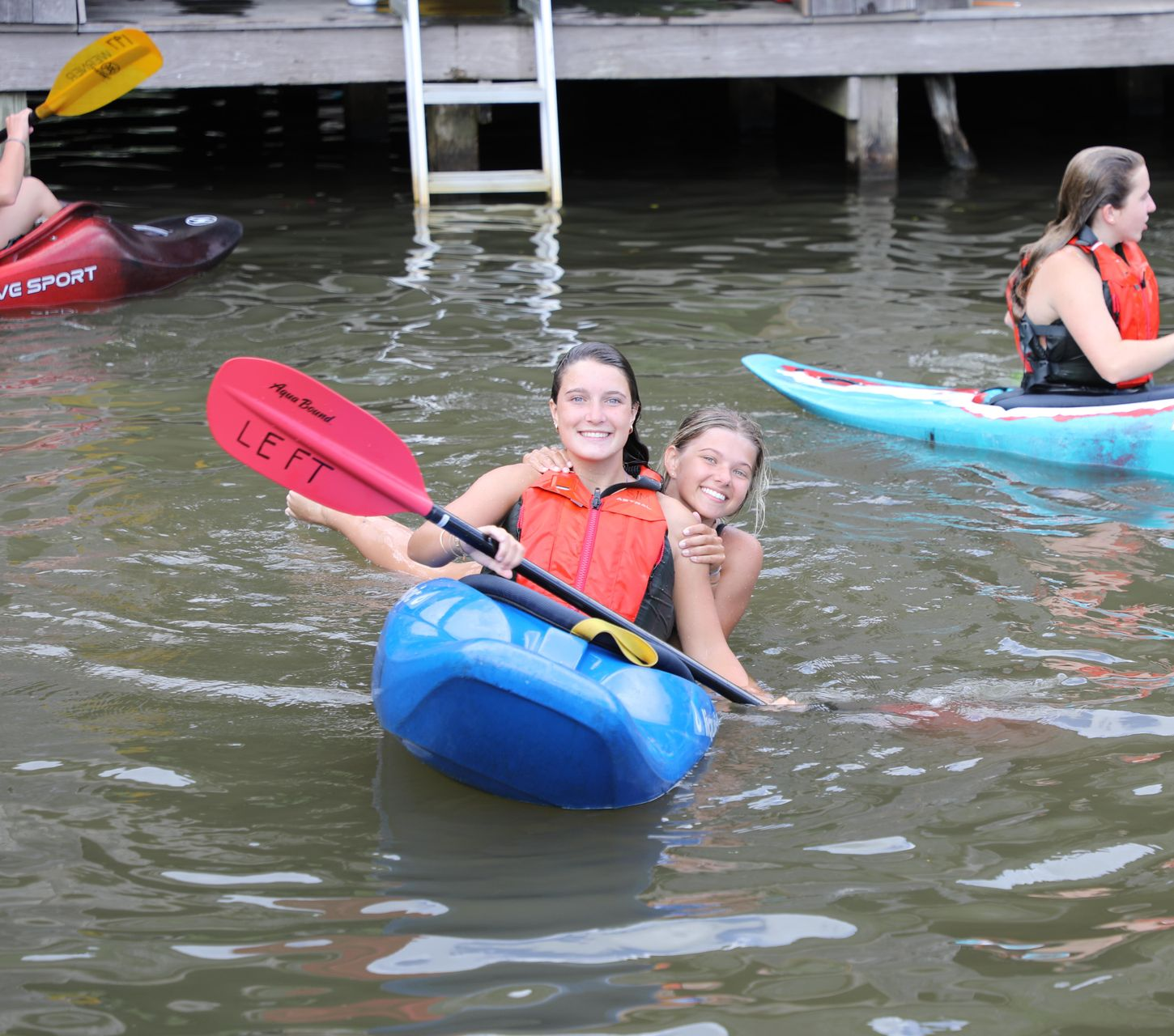 Two people share a blue kayak in a body of water, holding a red paddle with the word