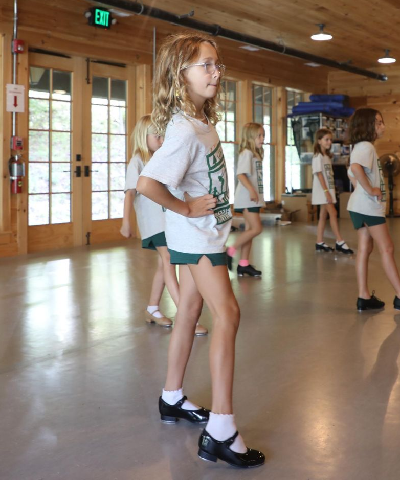 Dancers in white t-shirts and green shorts practice in a studio with wood walls and reflective floors.