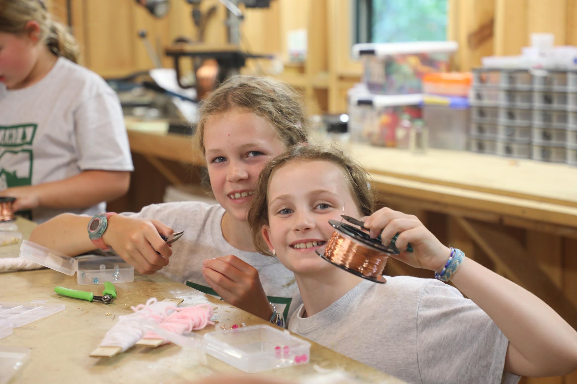 Two children smiling at a wooden workshop table, holding a spool of copper wire and jewelry-making tools.