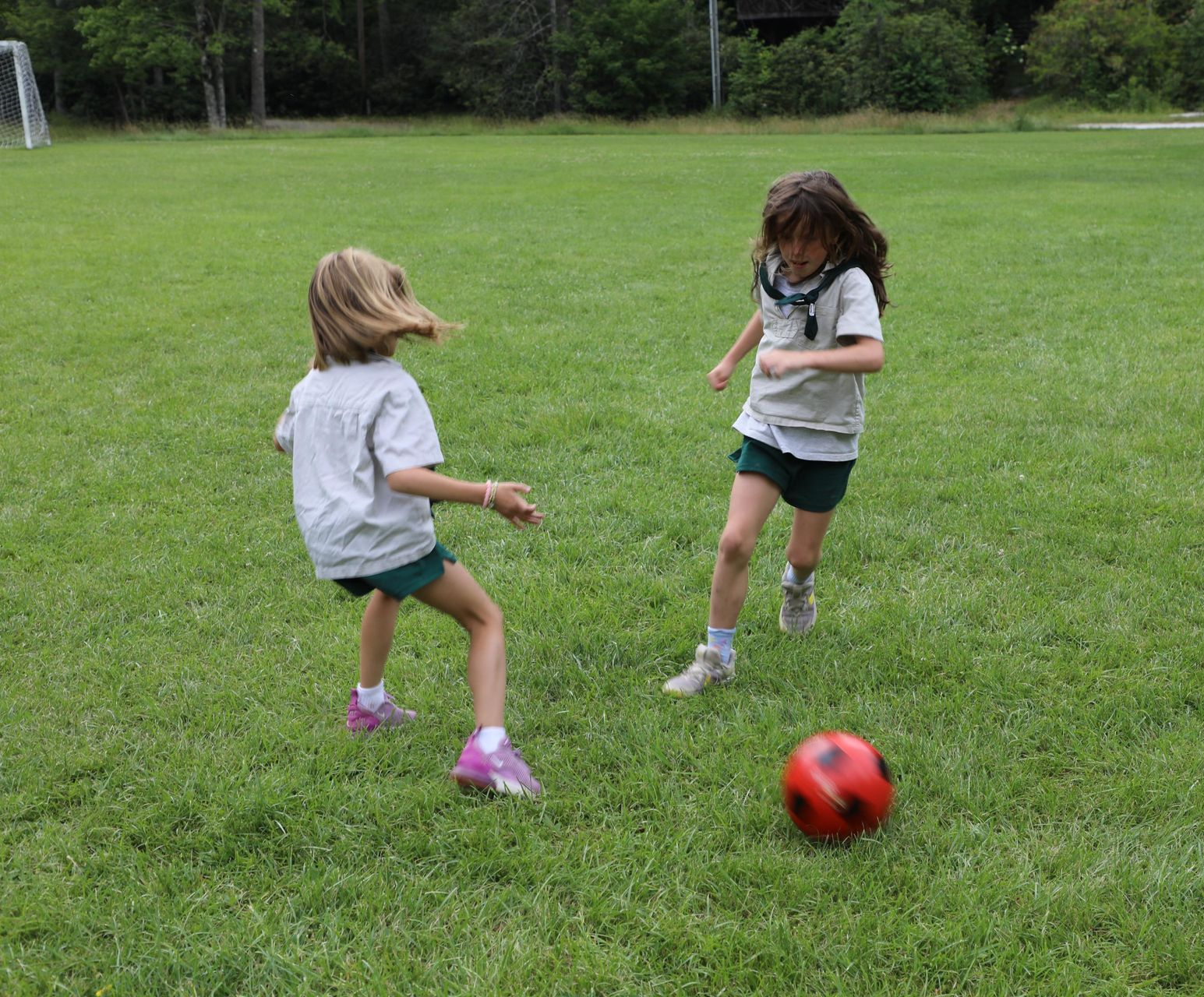 Two children in uniforms play soccer on a green grass field with a red ball.