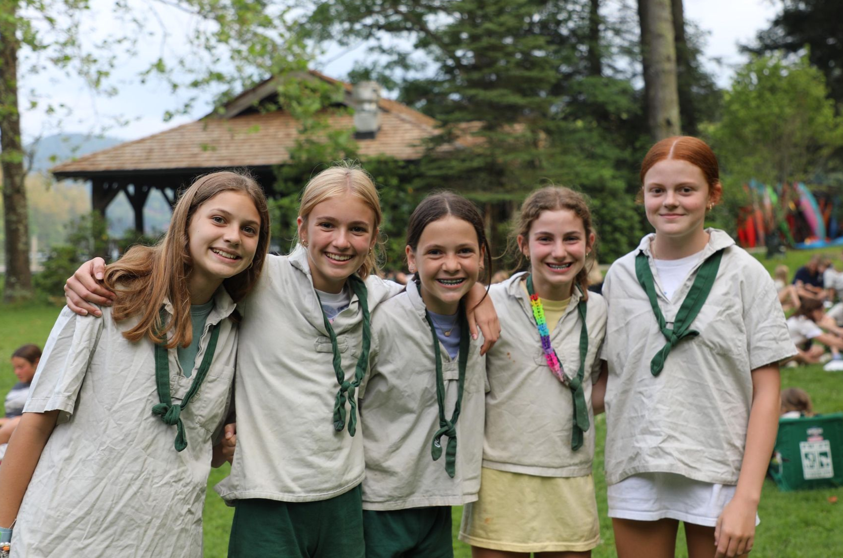 Five people in matching shirts and green scarves smiling while posing outdoors in front of a wooden building.