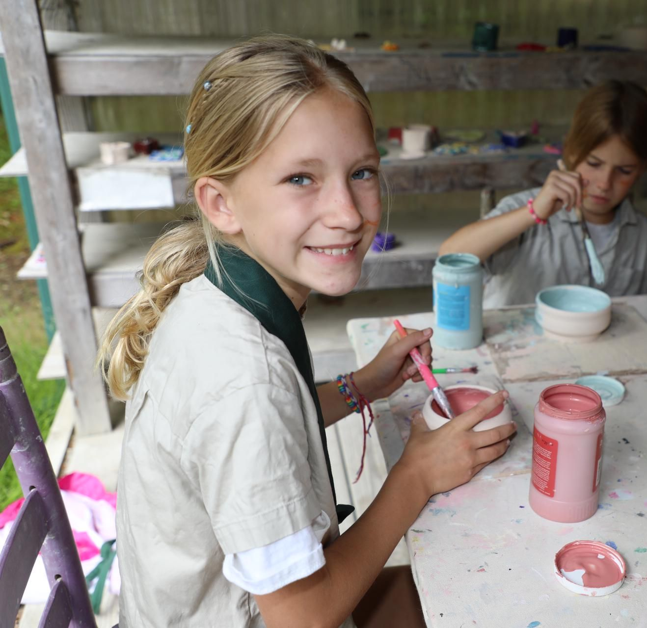 Two people wearing beige shirts sit at a table painting ceramic bowls with pink and blue paint in a workshop setting.