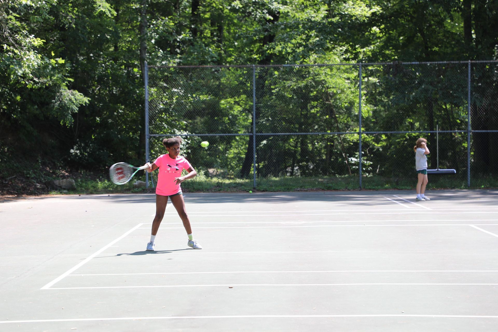 A person in a bright pink shirt swings a tennis racket toward a ball on an outdoor court, with another person in the back.