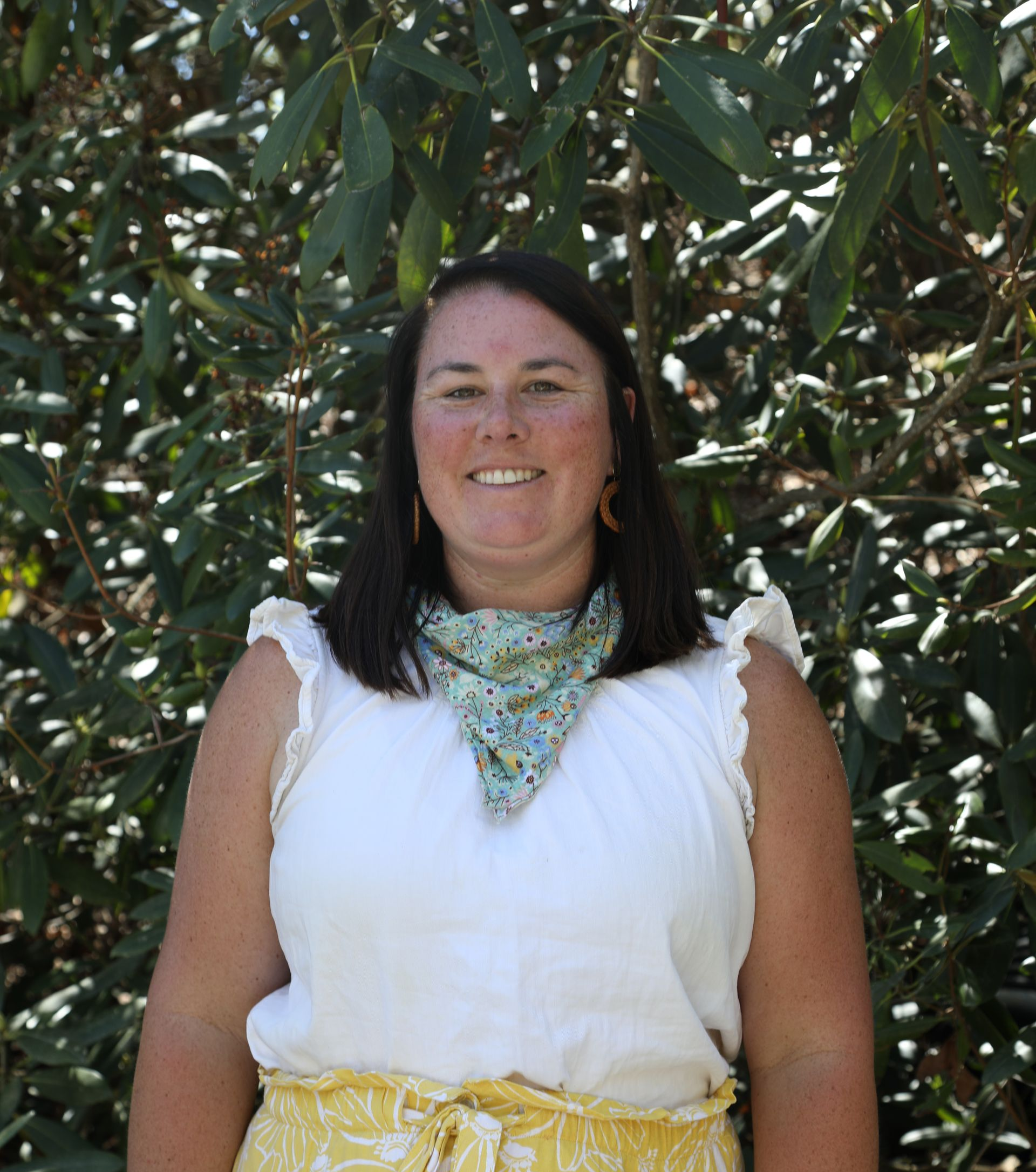 A person smiles while wearing a patterned neck scarf, a black shirt, and a light blue open button-down shirt outdoors.