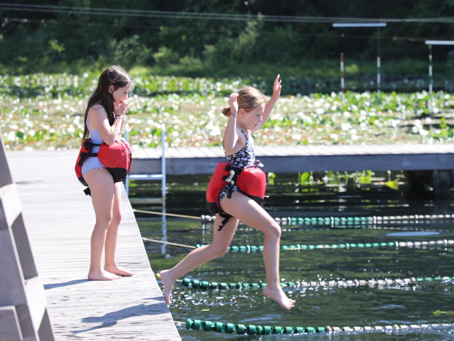 Two children in life jackets on a wooden dock, one standing and watching while the other jumps into the water.