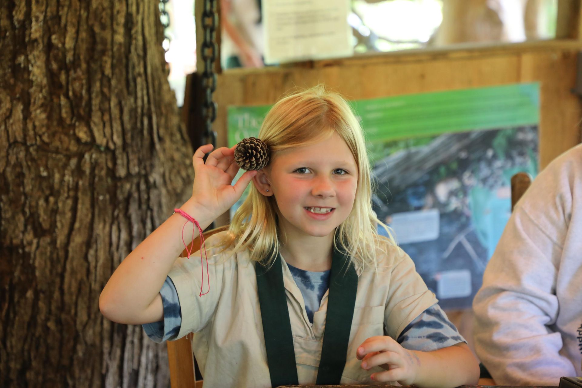 A person with blonde hair smiling while holding a pinecone near their ear in a rustic, outdoor setting.