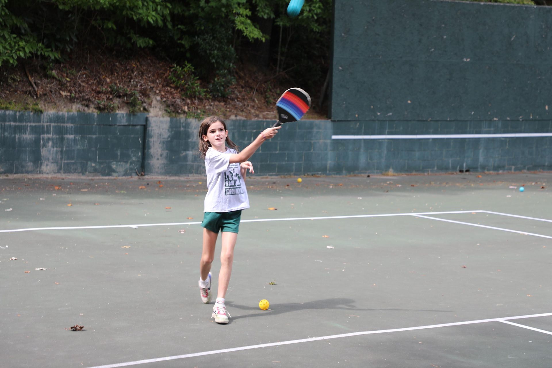 A young person in a white t-shirt and green shorts hits a ball with a colorful paddle on an outdoor court.