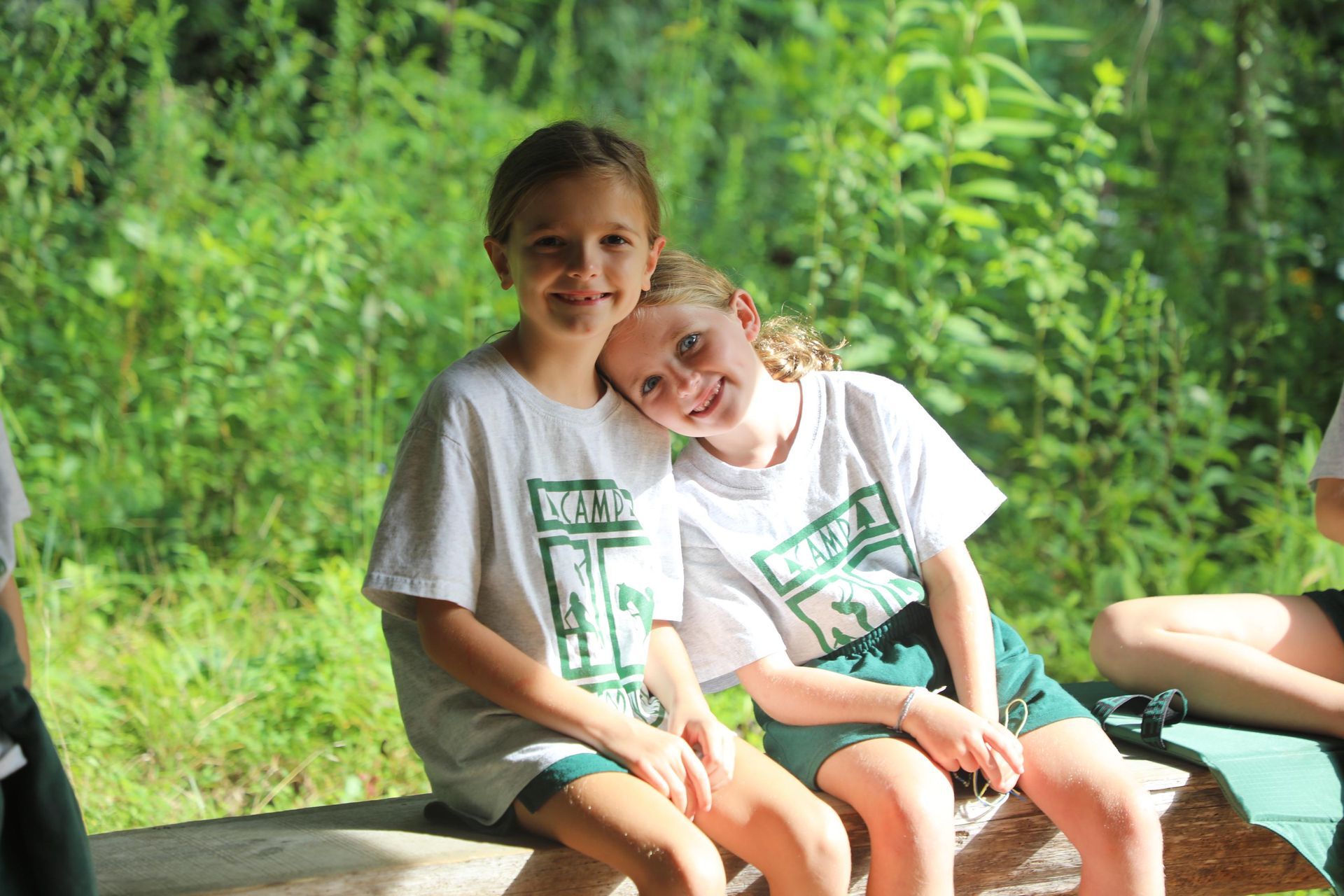 Two children in matching white t-shirts and green shorts sit side-by-side on a wooden log in a sunny, wooded area.