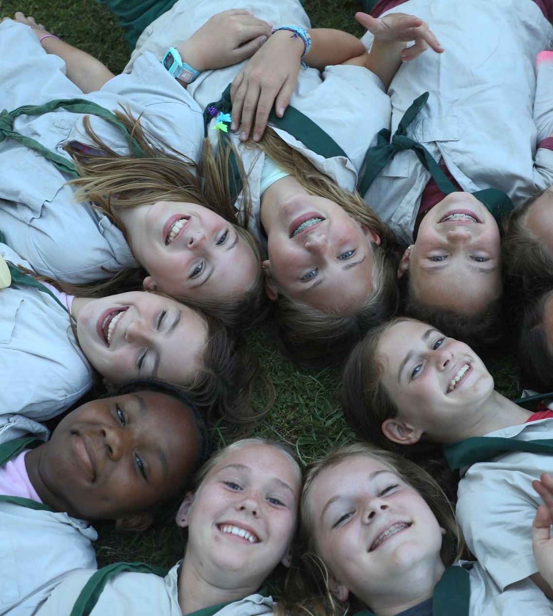 A group of people wearing uniforms and scarves lie in a circle on the grass, looking up and smiling at the camera.