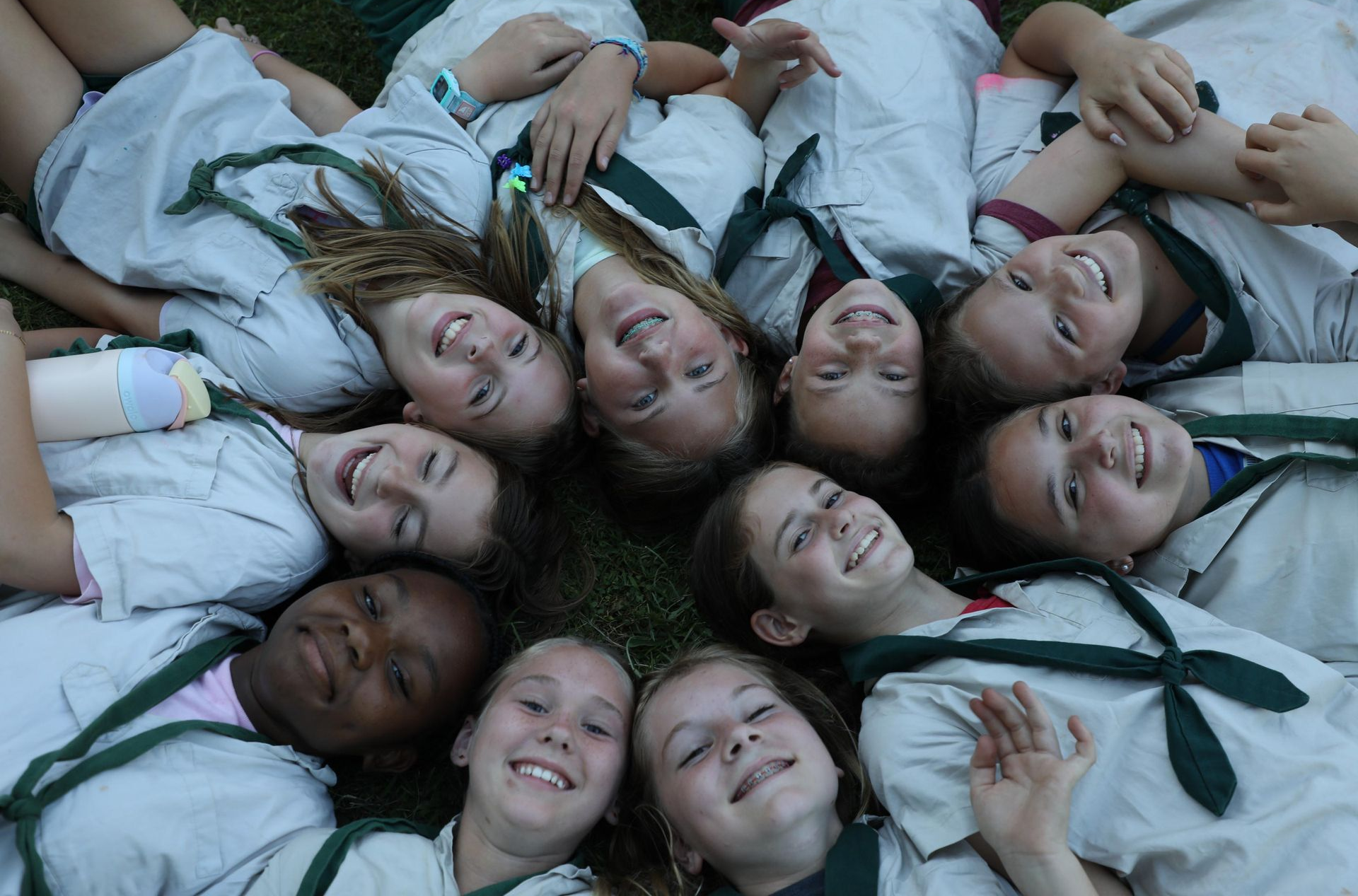 A group of scouts in tan uniforms with green neckerchiefs lying in a circle on grass, smiling up at the camera.