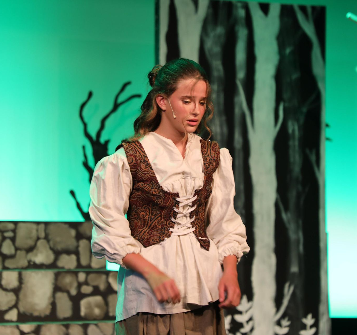 A person in a period costume with a lace-up bodice and white shirt performs on a stage with a painted forest backdrop.