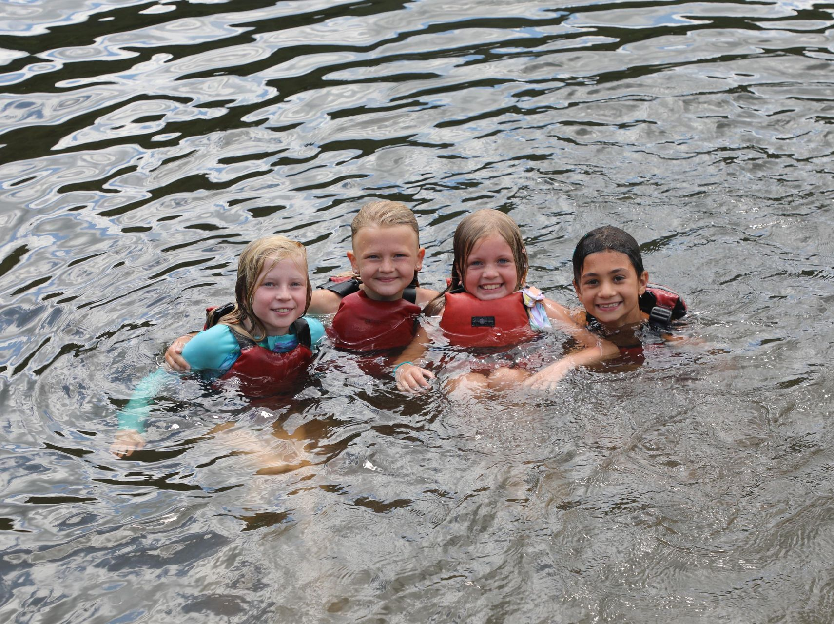 Four people wearing life vests smile while swimming together in a lake.