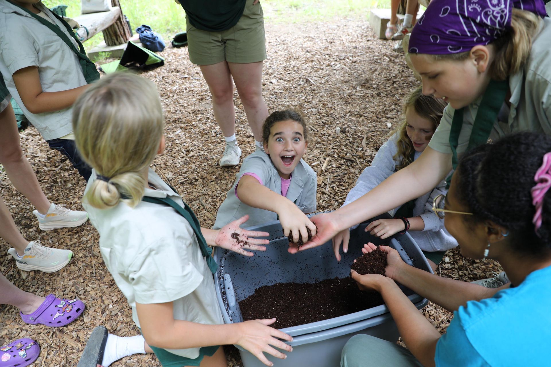 Group of children in matching uniforms gathering around a bin of soil, one child looking up with an excited expression.