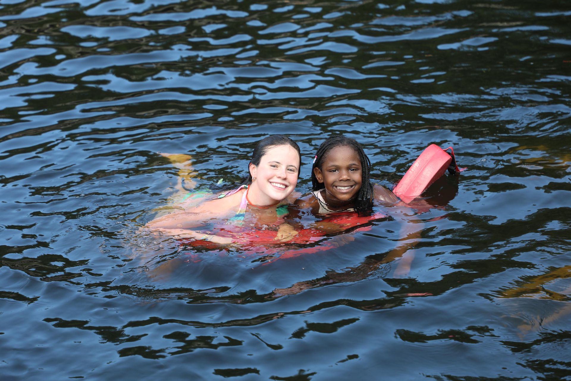 Two people smiling while swimming together in dark water, holding onto a red floating device.