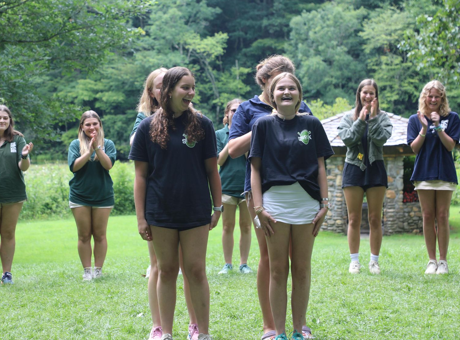 A group of people in matching uniforms standing on a grassy field in front of a stone structure, some clapping.