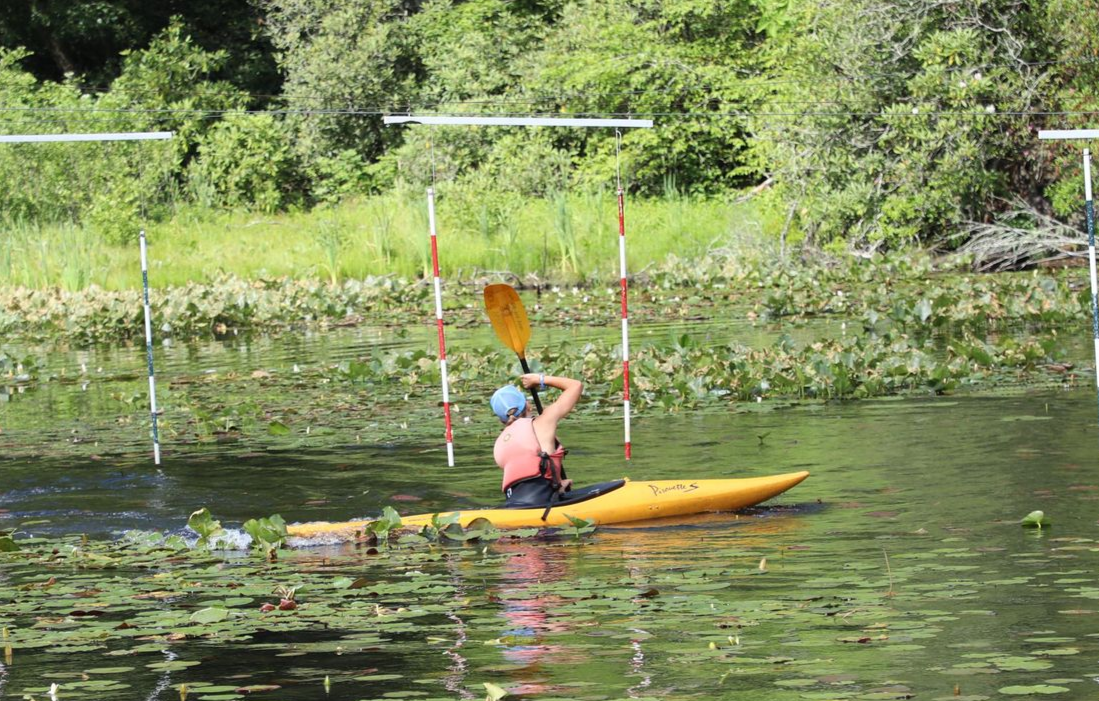 A kayaker in a life vest paddles a yellow kayak through a water course marked by red and white poles on a lily-pad pond.