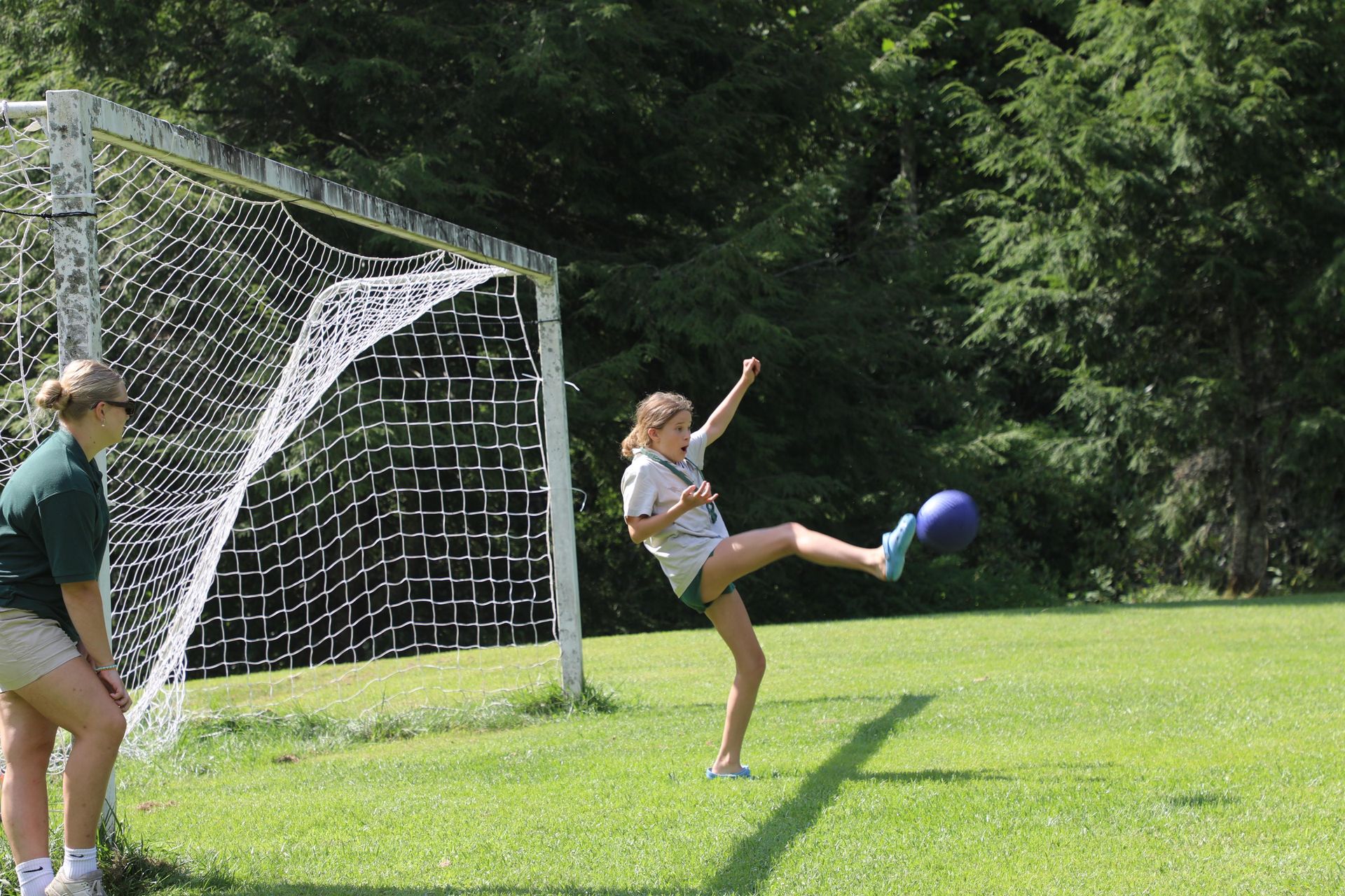 A person kicks a purple ball toward a soccer goal while another individual watches from the side on a grassy field.