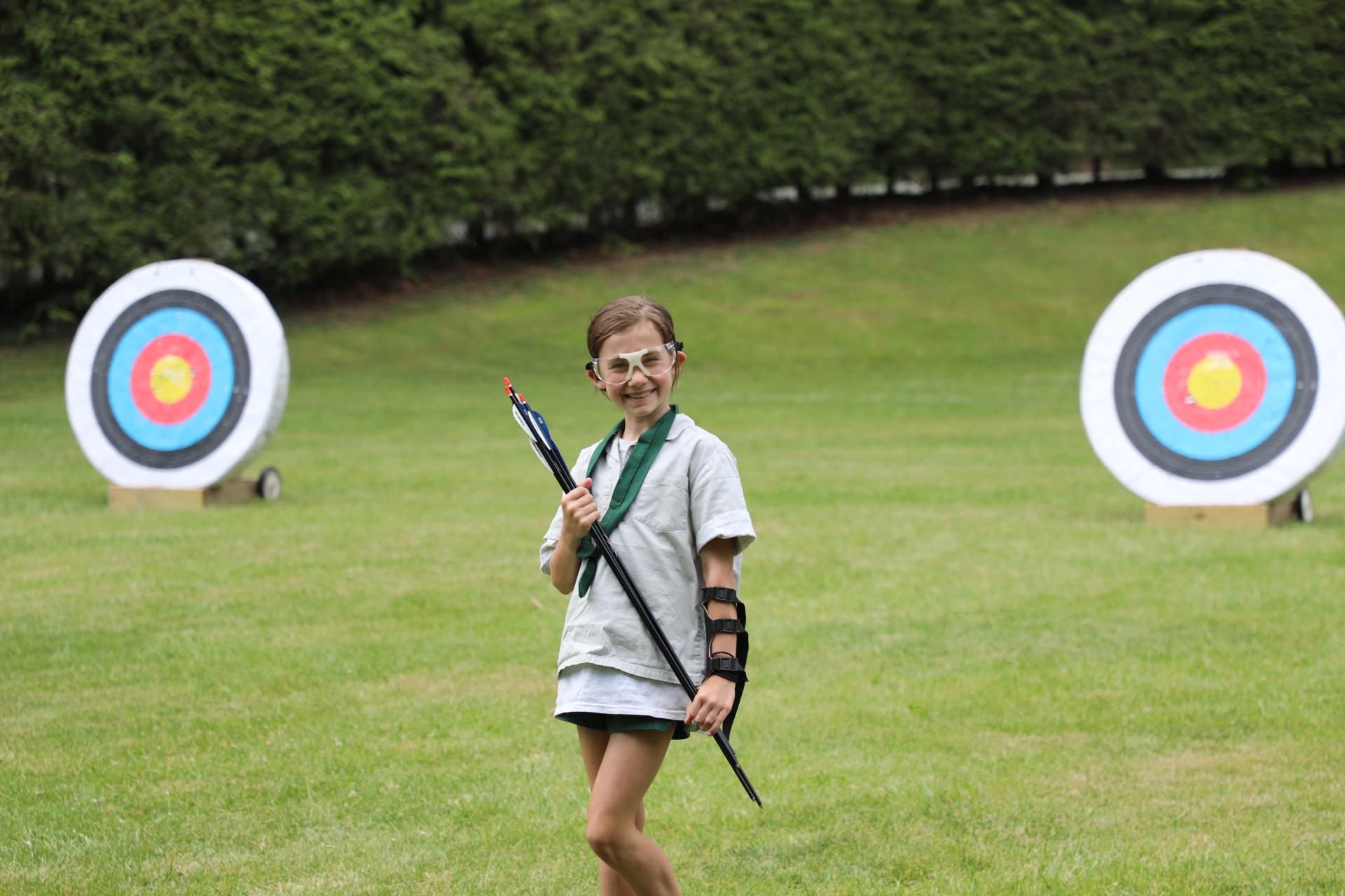 A smiling person holds a bow and wears an arm guard, standing on a grassy field between two archery targets.