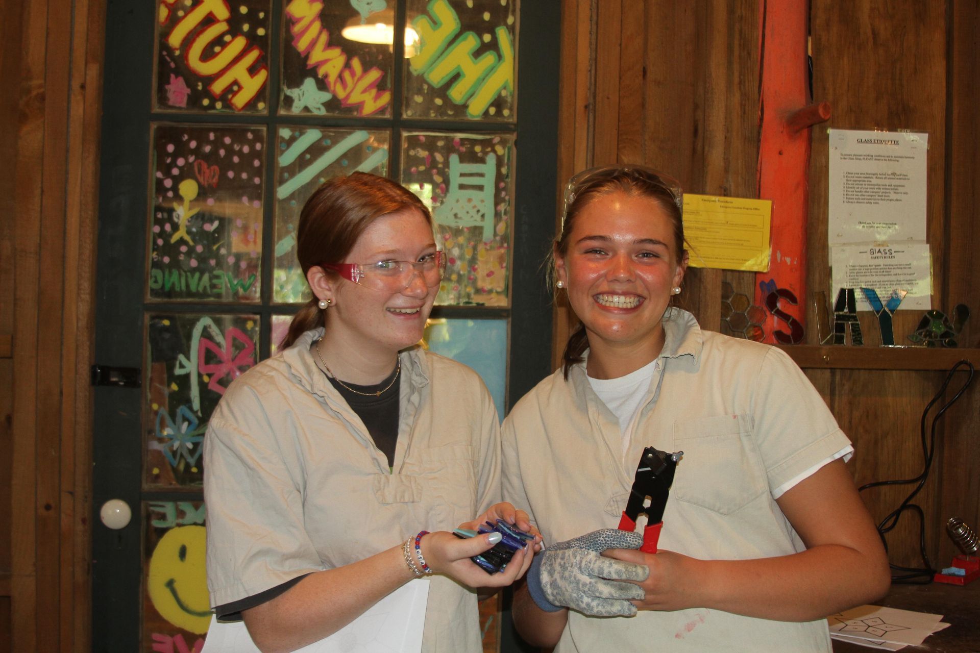 Two people in work shirts smiling while holding tools in a workshop with a decorated glass door in the background.