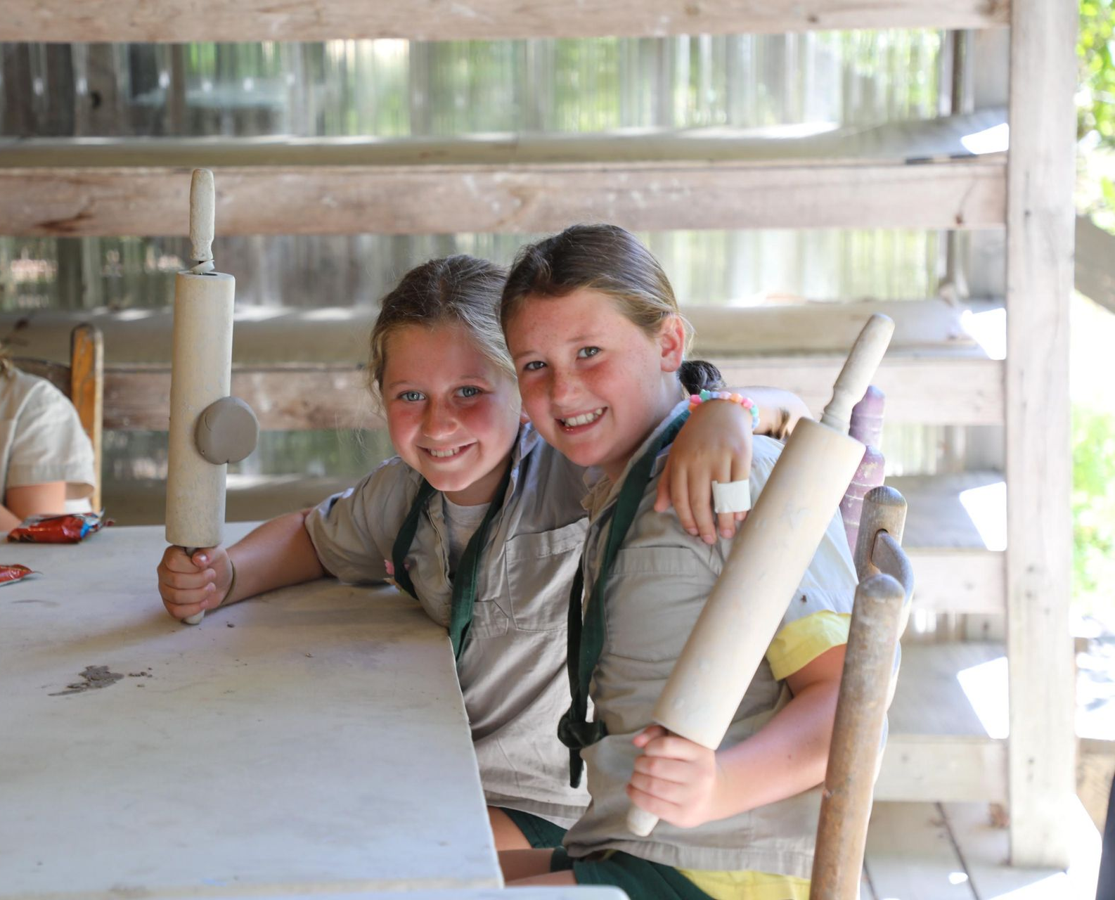 Two children in tan shirts smiling while sitting at a table and holding wooden rolling pins in an outdoor workshop.