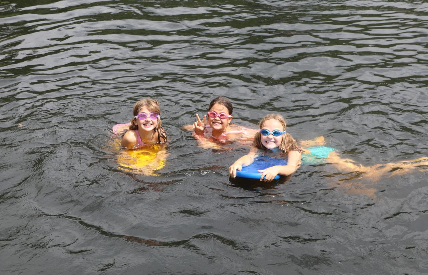 Three children wearing swim goggles swim in a lake using colorful kickboards.