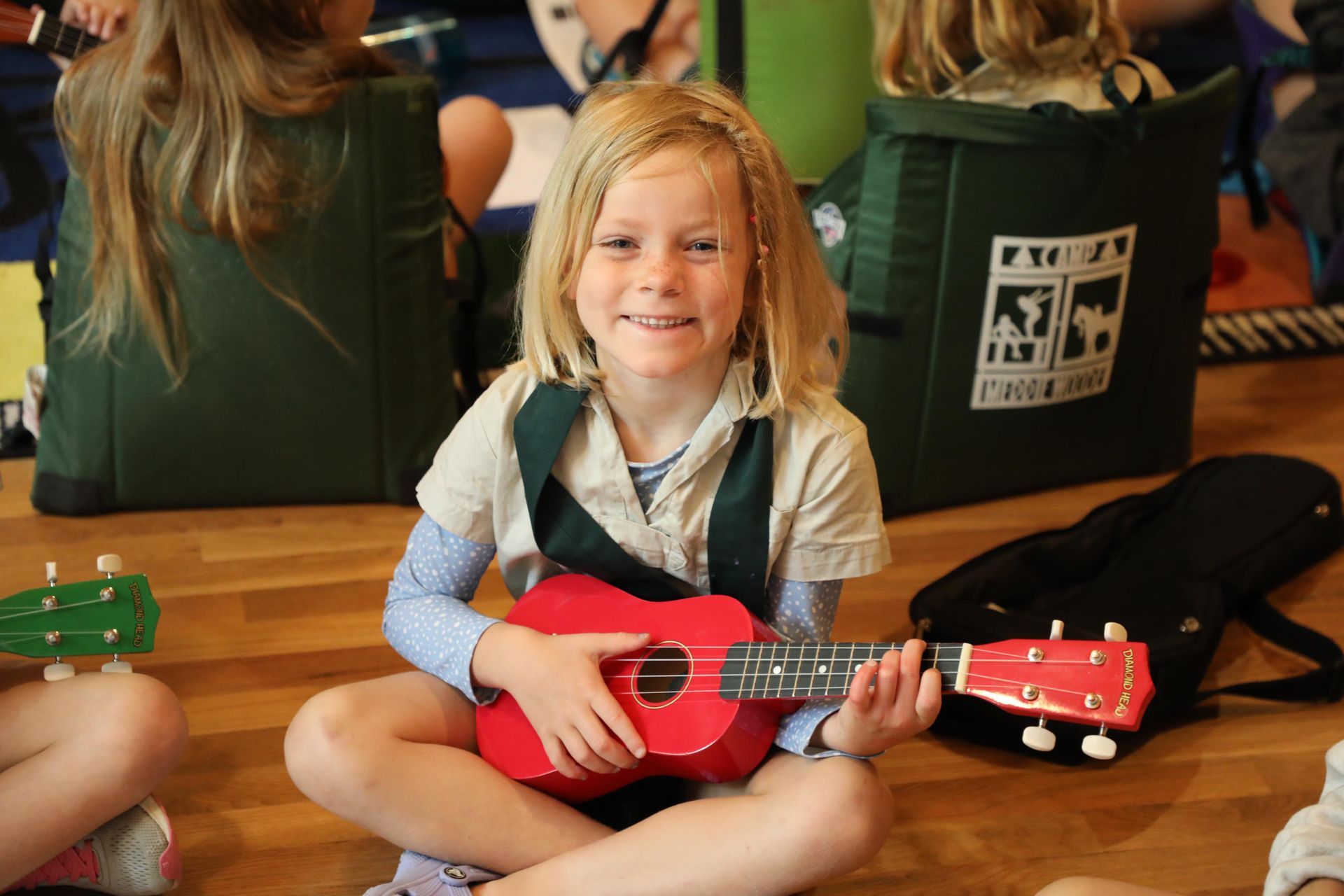 A smiling child sits cross-legged on a wooden floor, holding a small red ukulele while participating in a music class.