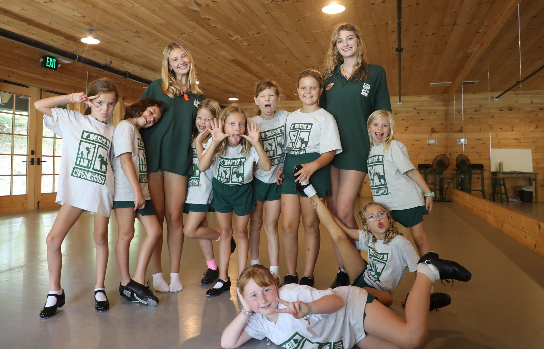 A group of individuals in matching t-shirts pose for a photo in a wooden studio, with one lying on the floor in front.