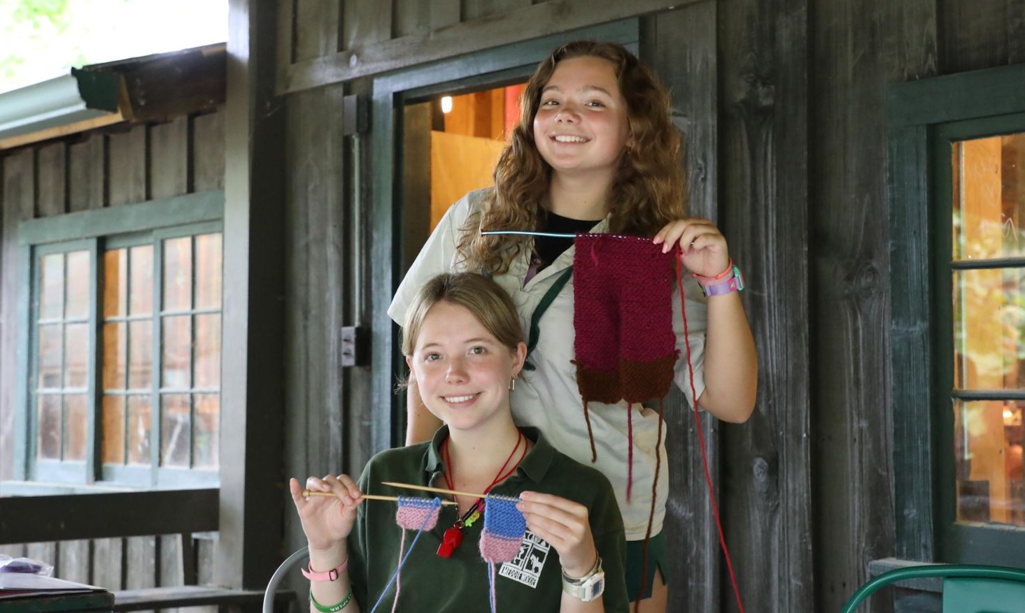 Two people on a wooden porch smiling while holding knitting projects with needles and yarn.