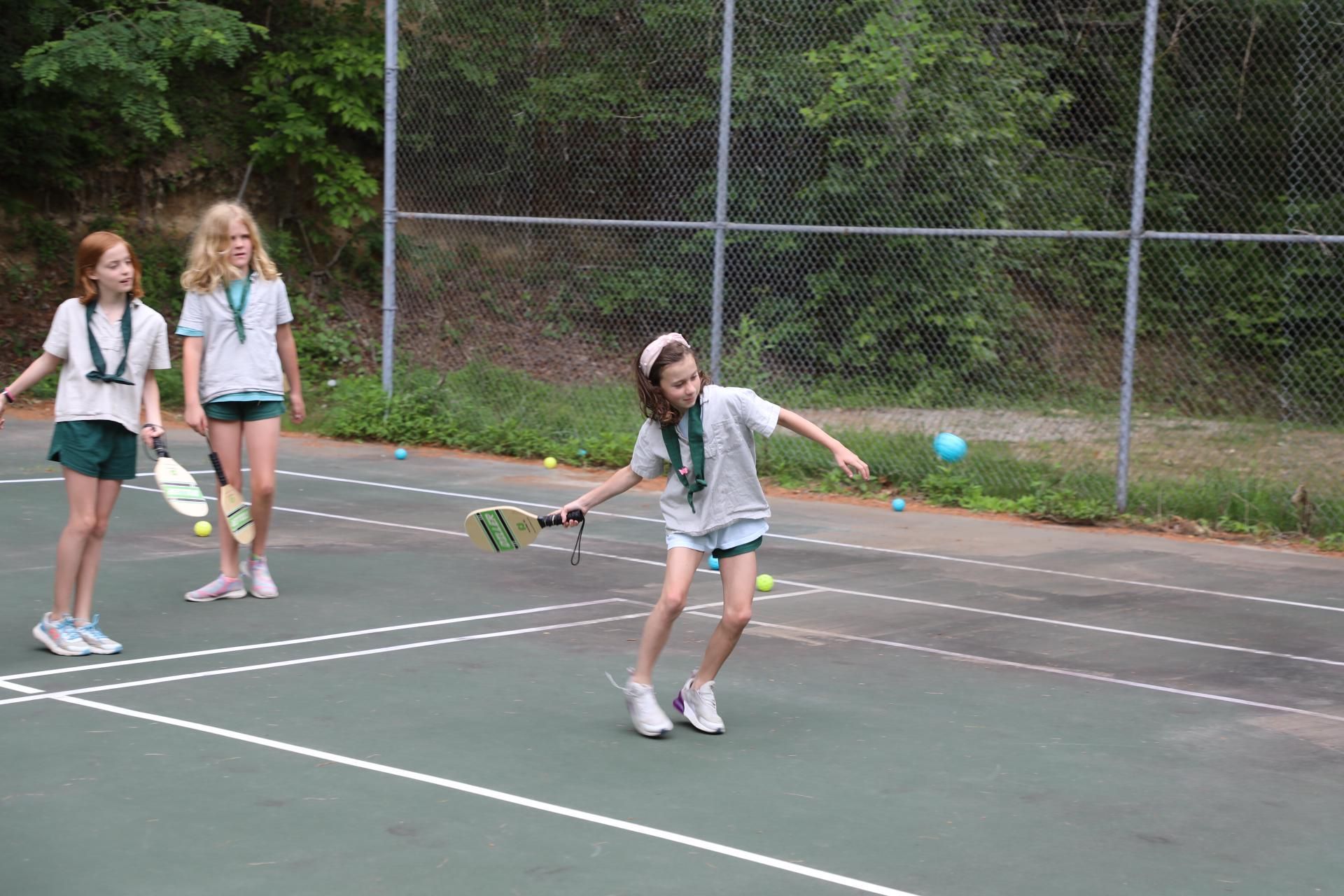 Three people play pickleball on an outdoor green court surrounded by a chain-link fence.