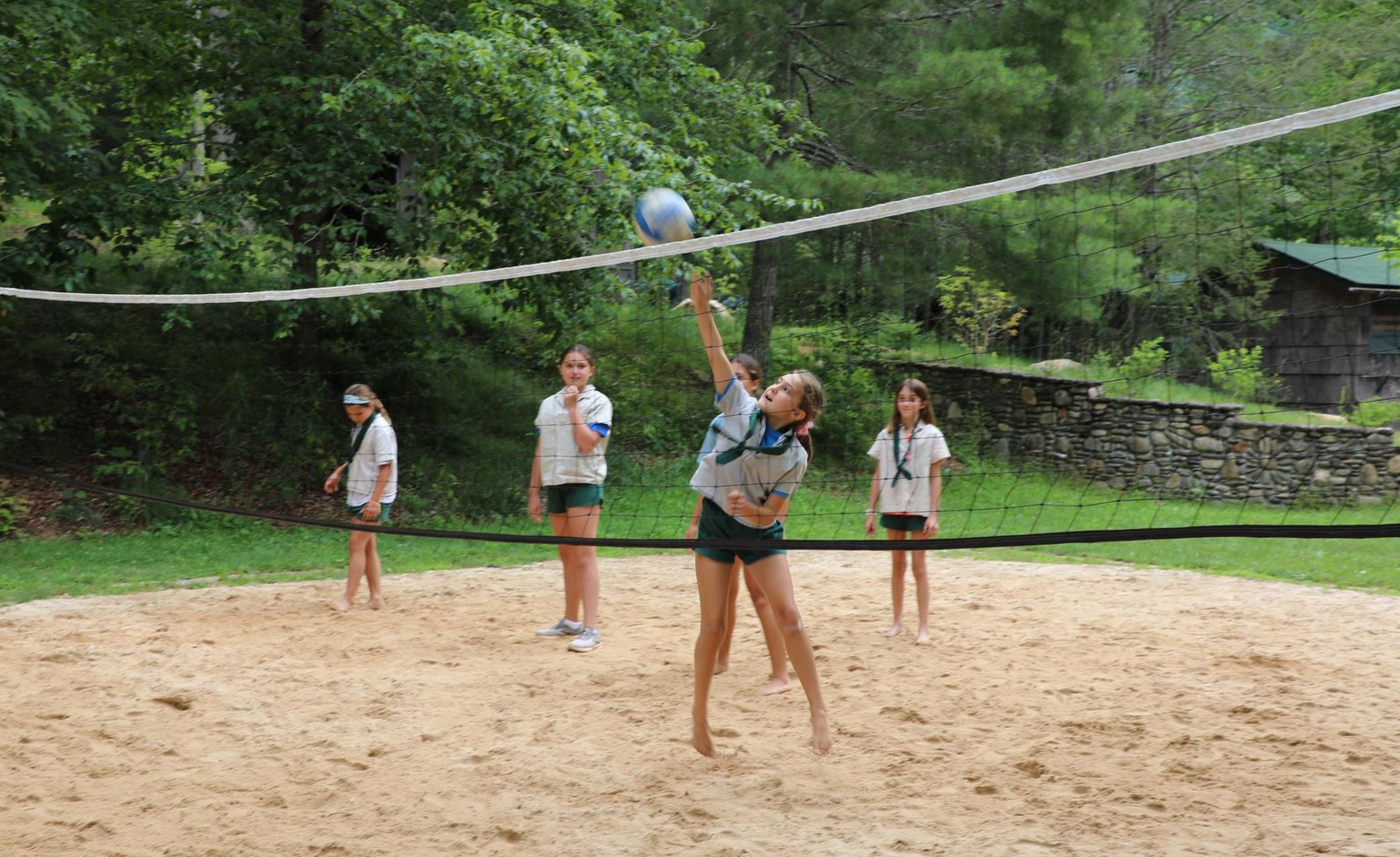 A group playing volleyball on a sand court with a net, one player jumping to hit the ball.