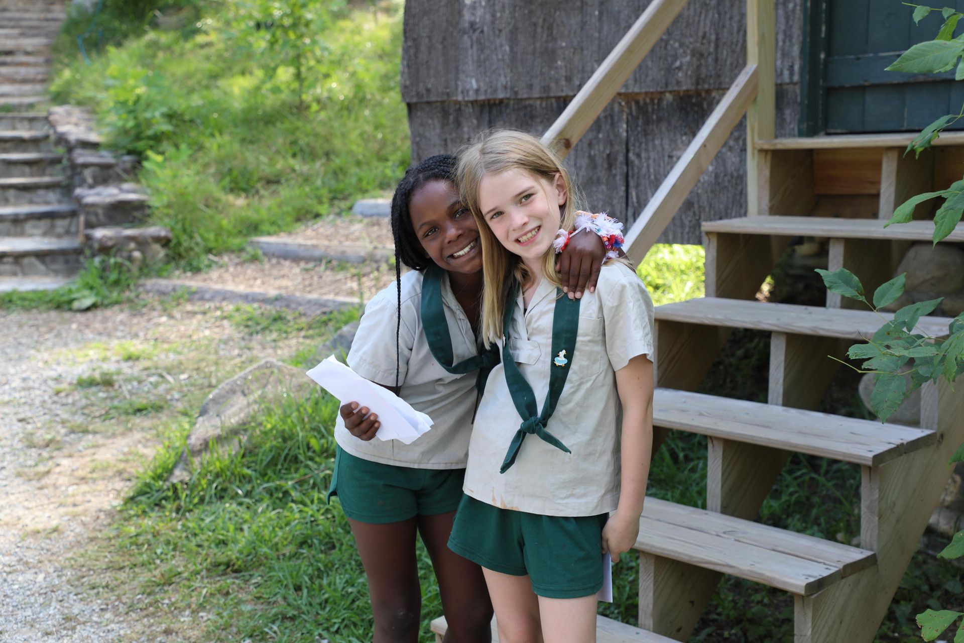 Two campers in tan uniform shirts and green shorts smiling with arms around each other next to wooden outdoor stairs.