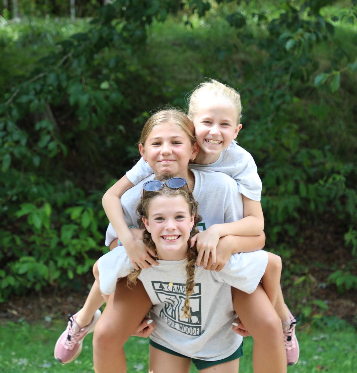 Three people smile while posing for a piggyback photo in front of a leafy green background.