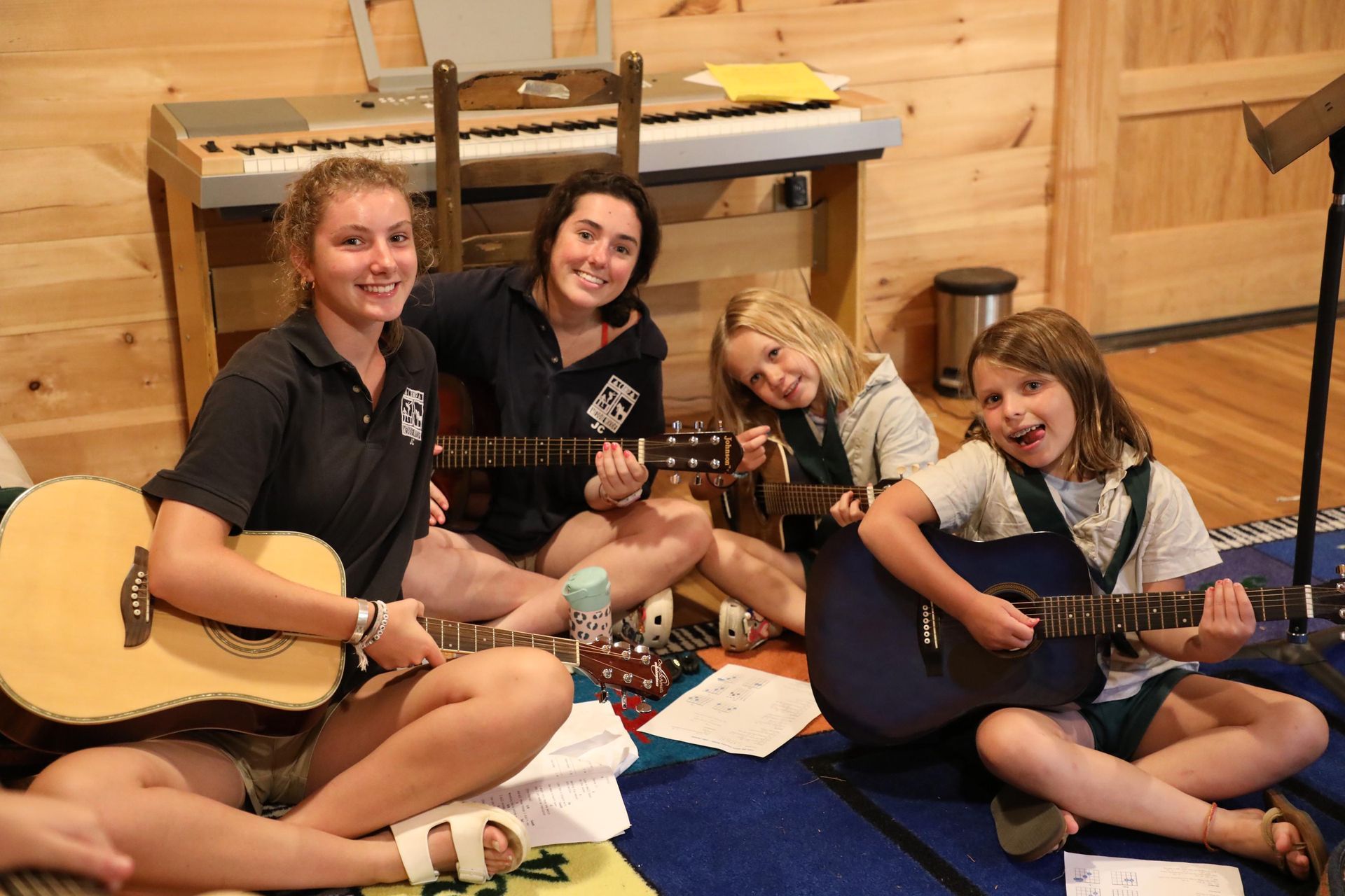 Four people sit on a blue rug in a wooden room, smiling while holding and playing acoustic guitars.