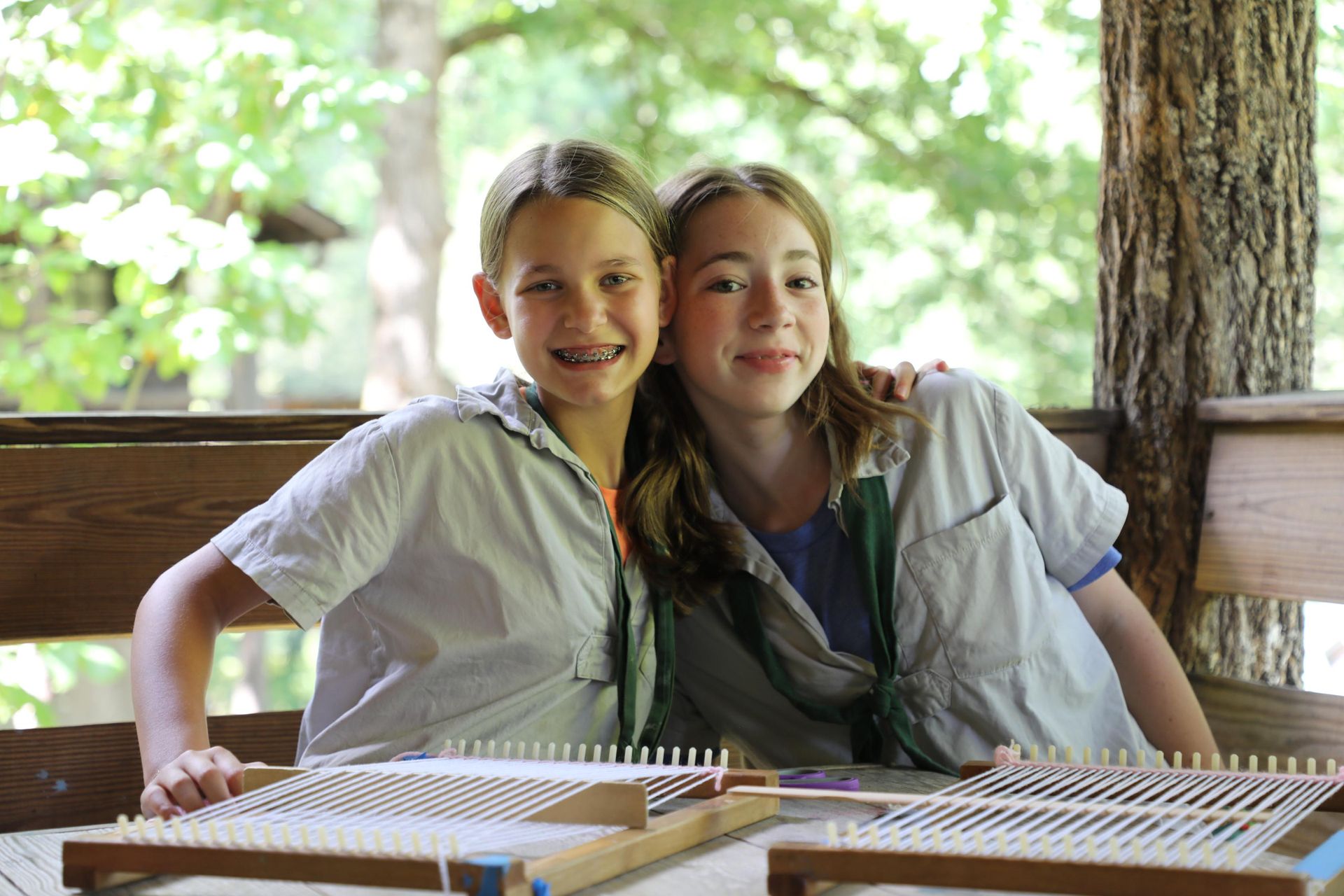Two people sit side-by-side on a wooden porch, smiling while working on individual hand looms with white woven thread.