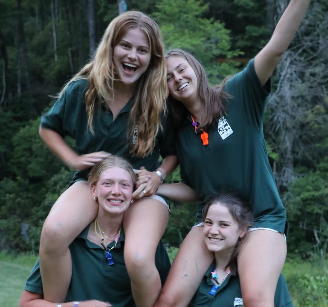 Four people in matching green shirts pose outdoors, two perched on the shoulders of the others, all smiling.