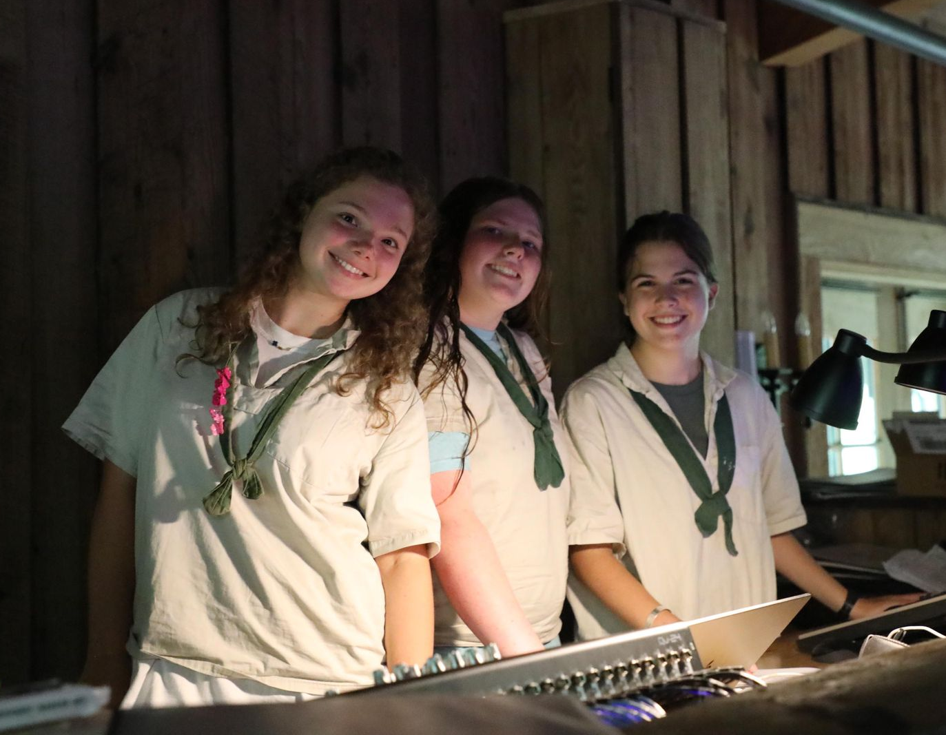 Three people in cream-colored shirts with green neckerchiefs smile while standing behind a sound mixing console.