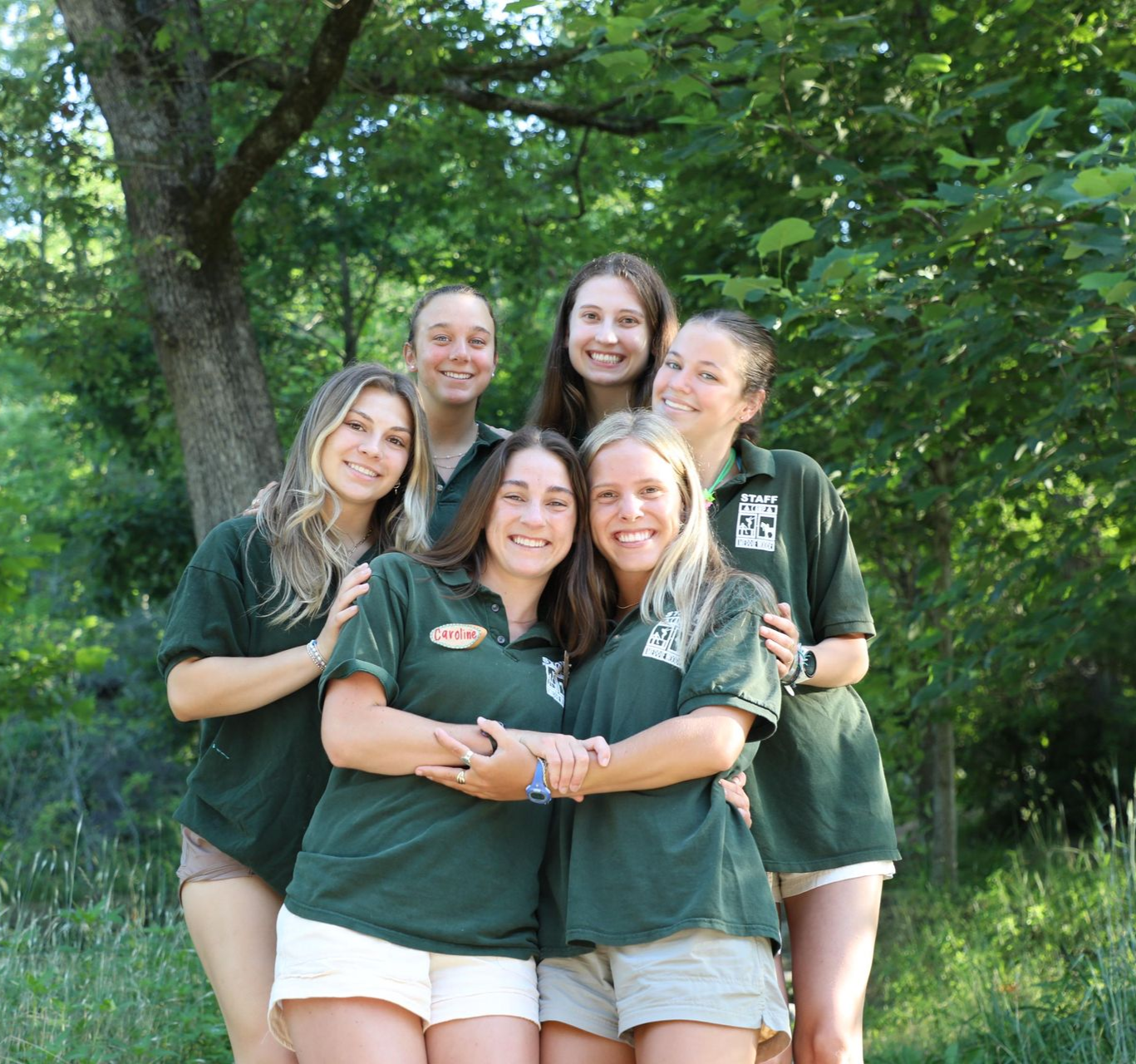 Six people smiling together outdoors in a lush, green wooded setting, wearing matching green polo shirts and shorts.