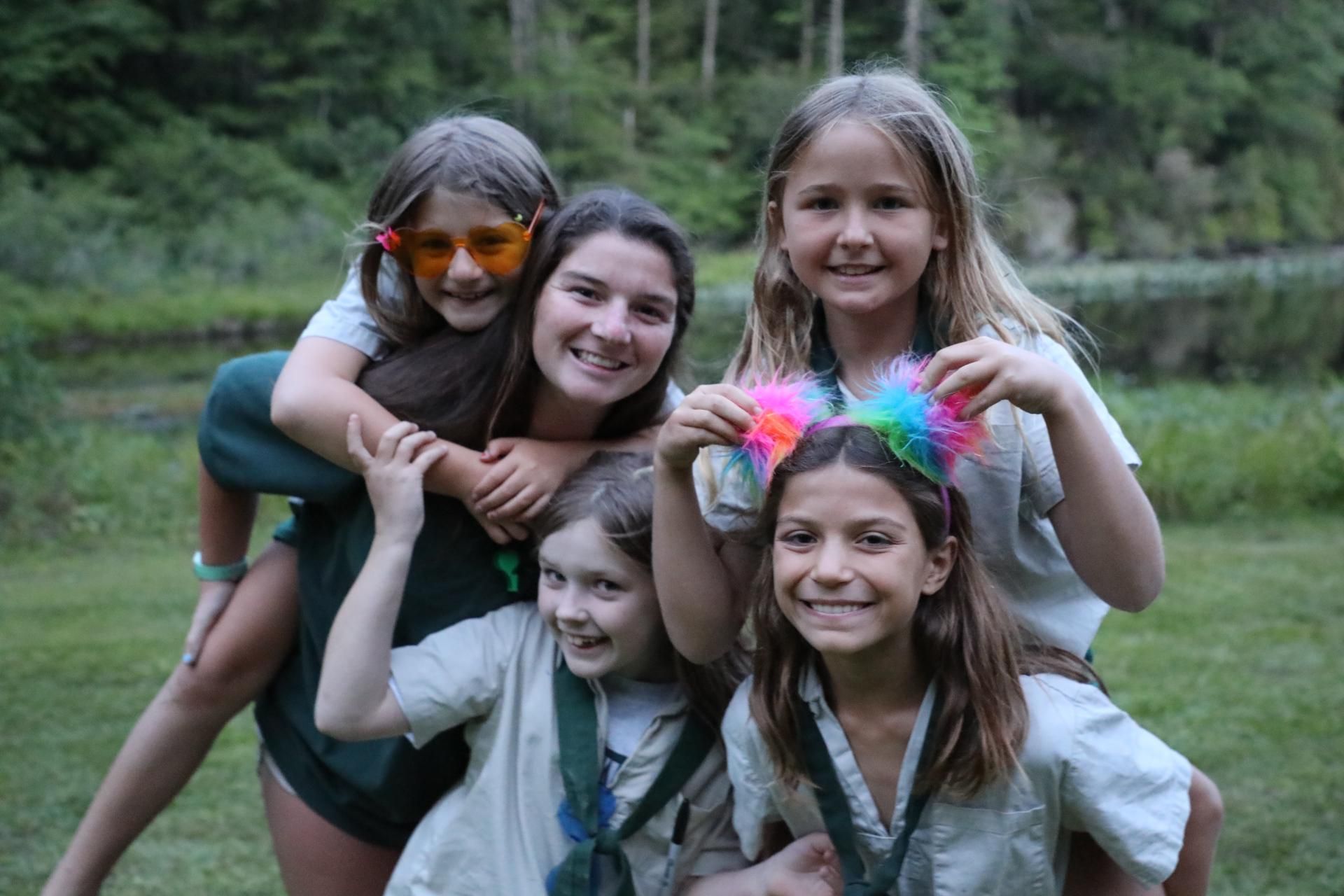 Five people in camping attire pose together outdoors, some smiling and one wearing a colorful fuzzy headband.