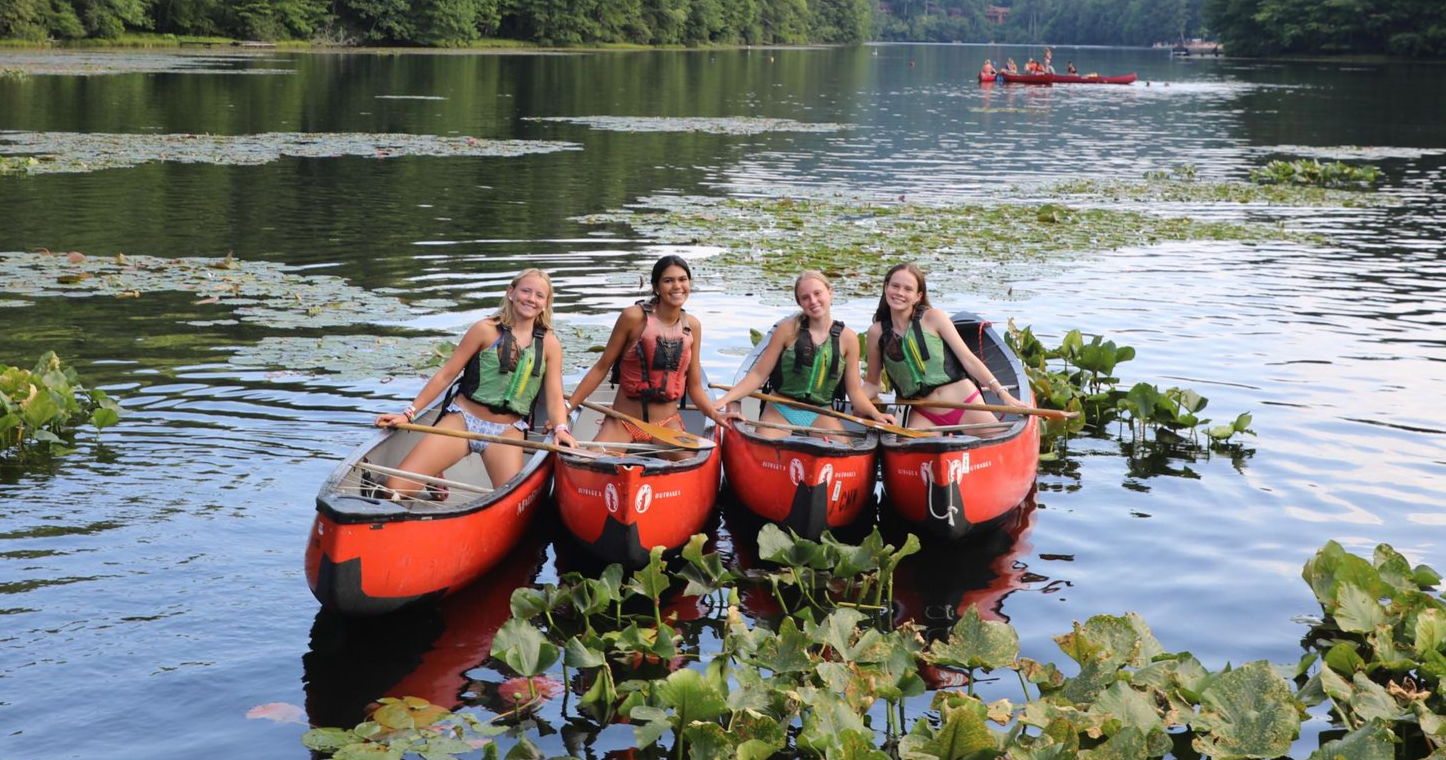 Four people sitting in two red canoes on a lake surrounded by lily pads, wearing life jackets and smiling.