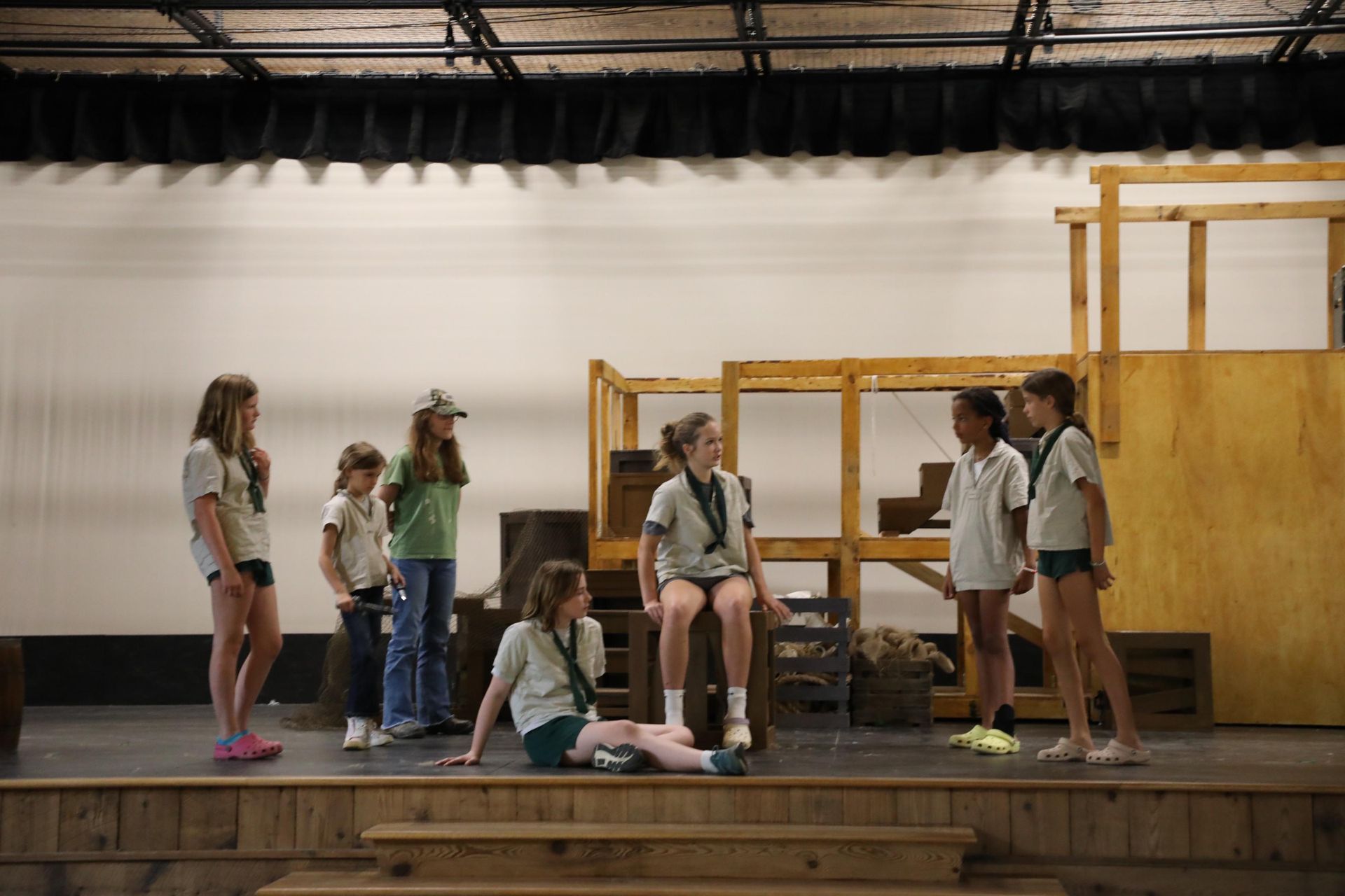 Six youths in casual clothing gather on a wooden stage with a wooden structure background, engaged in a conversation.
