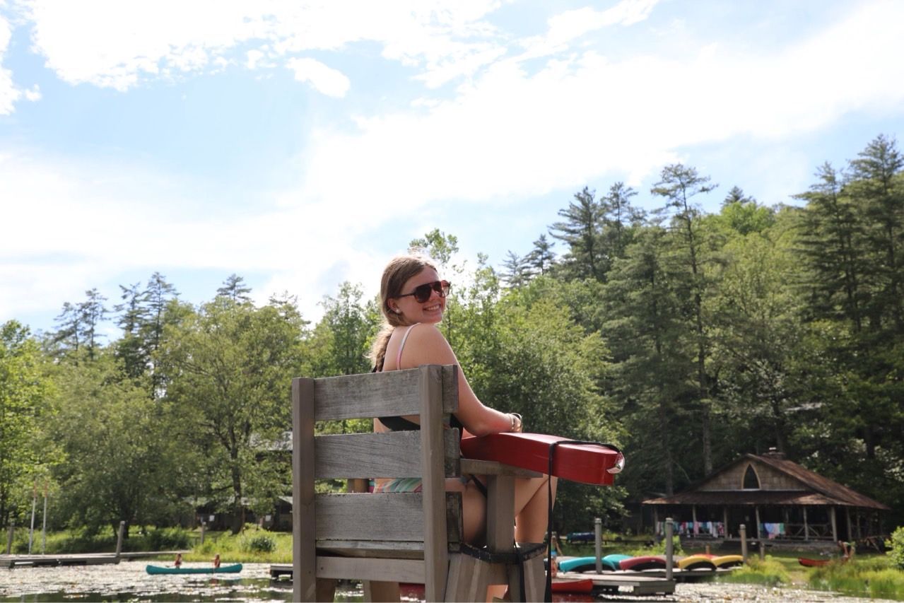 A lifeguard sits on a wooden high chair overlooking a sunny lake with a wooded shoreline and a small building.