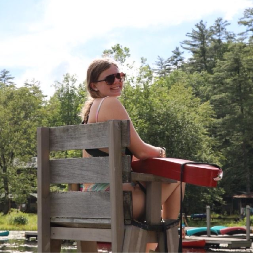 A lifeguard wearing sunglasses sits on a wooden high chair overlooking a sunny lake with trees and small boats nearby.