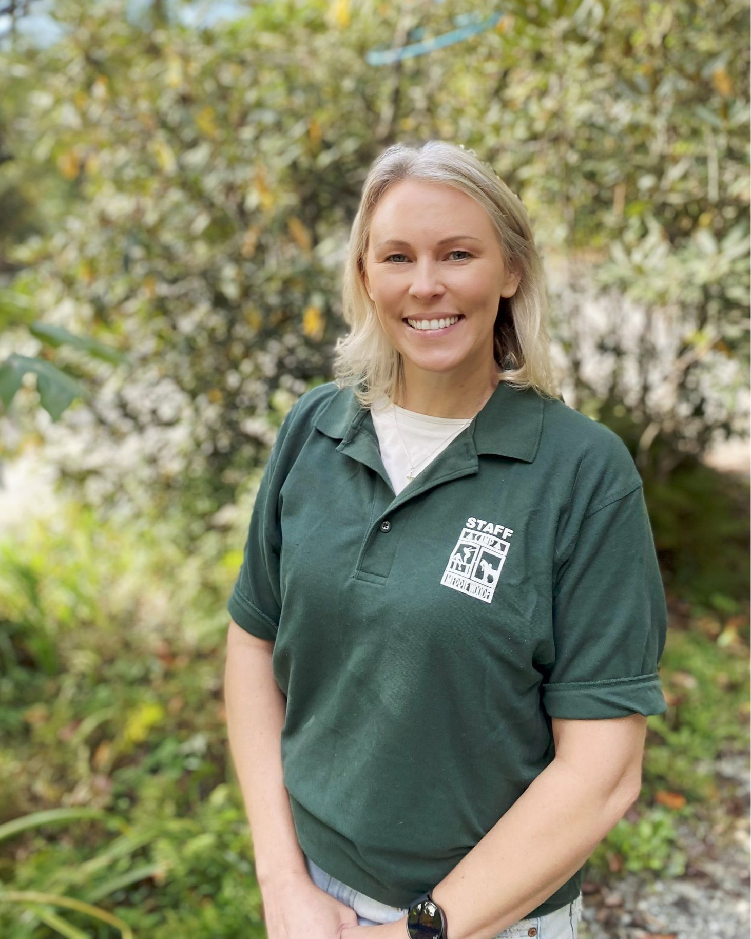 A smiling person wearing a dark green polo shirt stands outdoors in front of a leafy green bush.